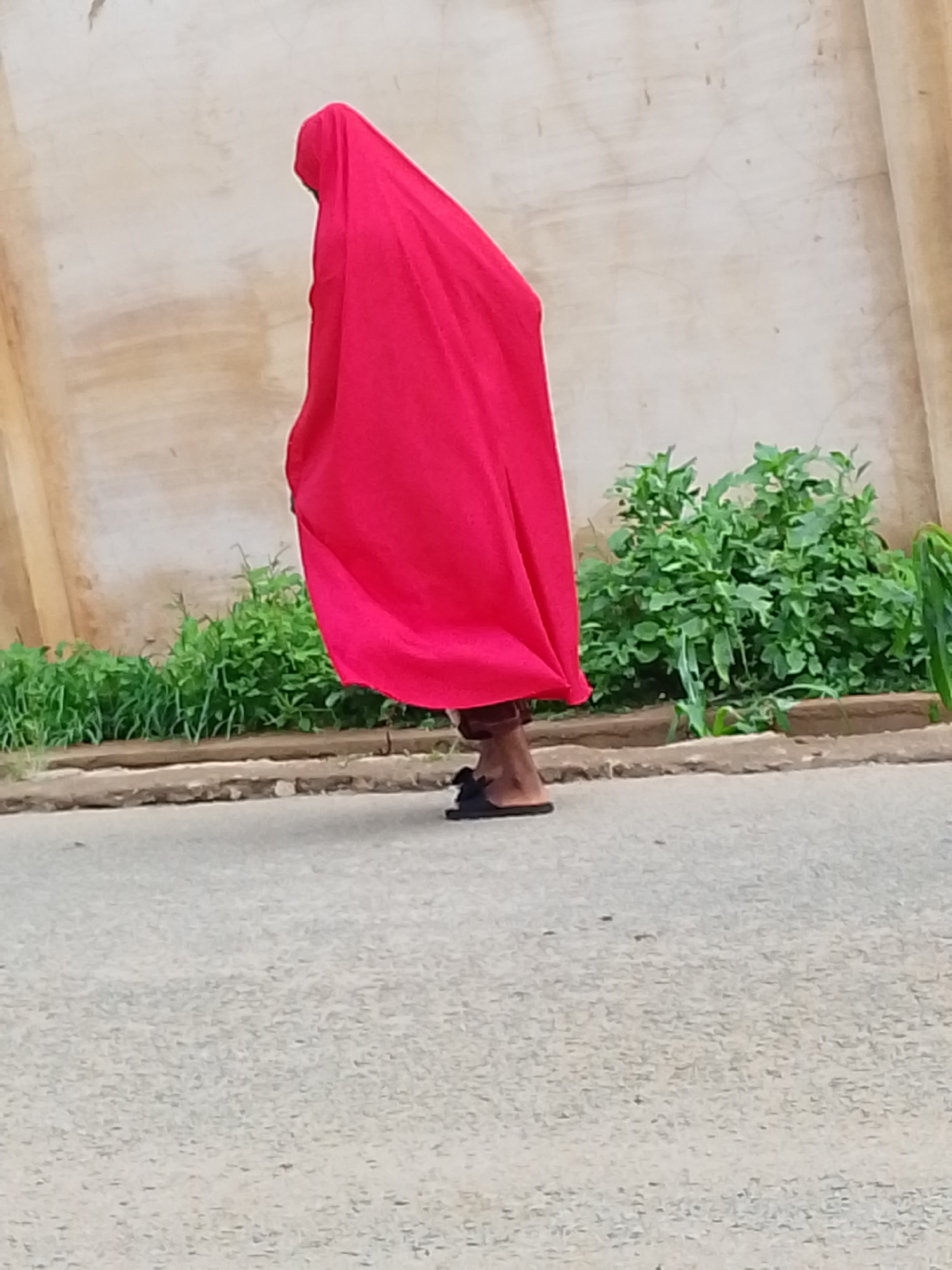 A person is draped in a red cloth, obscuring their identity and form. Only their feet, shod in black sandals, are visible. They stand on a paved surface, likely a road, in Jos, Nigeria. Behind them is a low wall bordering green plants and grass. A light-colored building forms the backdrop of the scene.