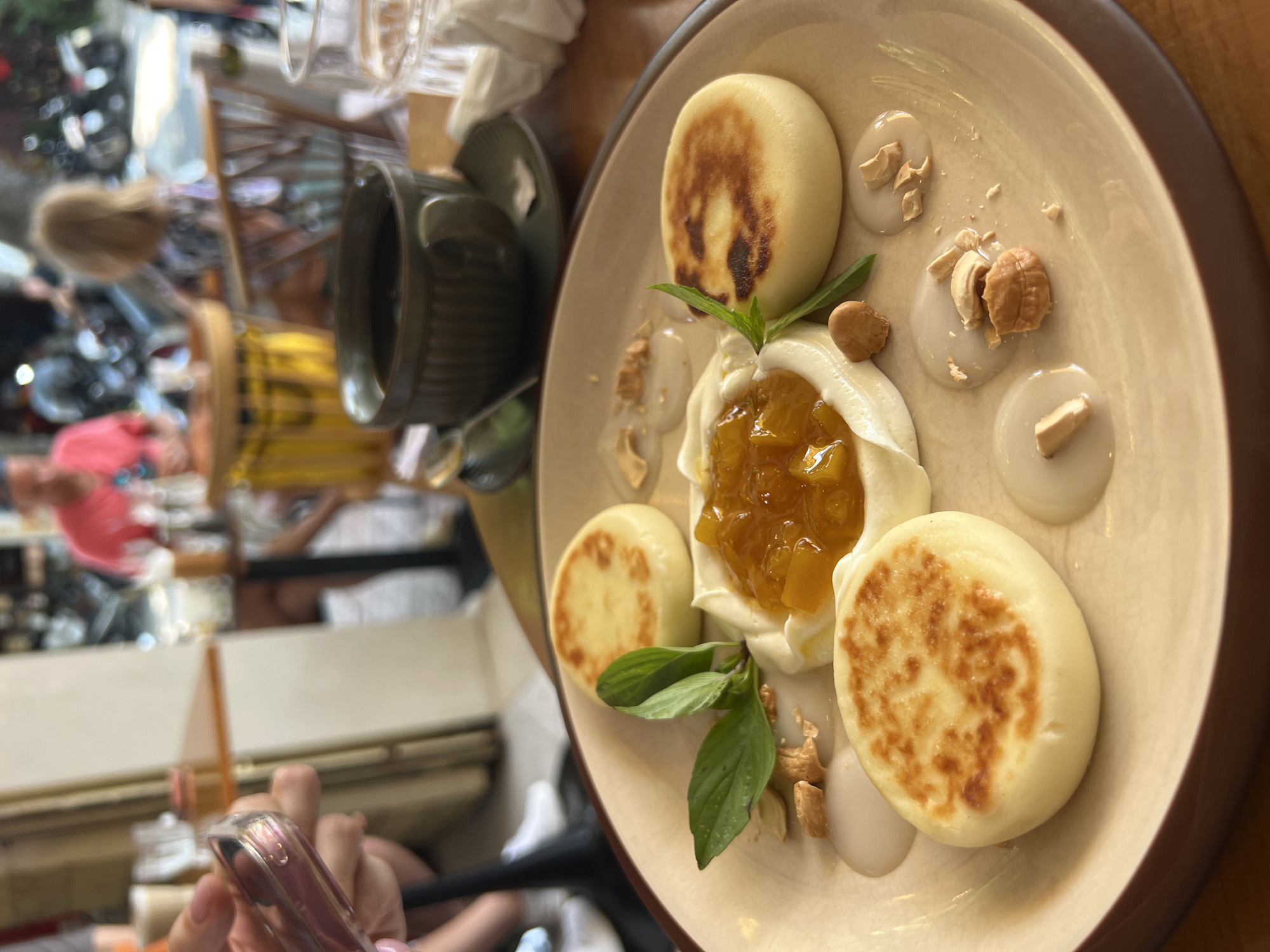 Here is a description of the image:
The image captures a close-up, high-angle shot of a plate of Syrniki, Russian fried quark pancakes. The Syrniki are arranged around a dollop of white cream, topped with a yellow jam and fresh green basil. The plate also contains cashew nuts and what seems to be a sweet sauce. A dark green coffee mug sits to the left of the plate. In the background, there are people sitting at tables and what appears to be a reflective surface. A hand holding a cell phone is in the foreground.
Without additional information, it is impossible to confirm the location of Nha Trang, Vietnam.