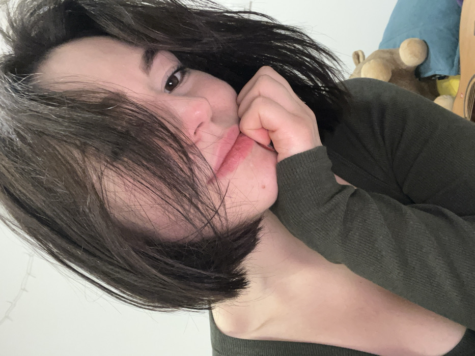 A young woman with dark, shoulder-length hair rests her face in her hand, looking at the camera. She wears a dark green long-sleeved shirt. Her lips are slightly parted, and her expression is soft. A mole is visible on her cheek. In the blurred background, there is a blue pillow, a teddy bear, and a guitar. The photograph is taken at a slightly skewed angle.