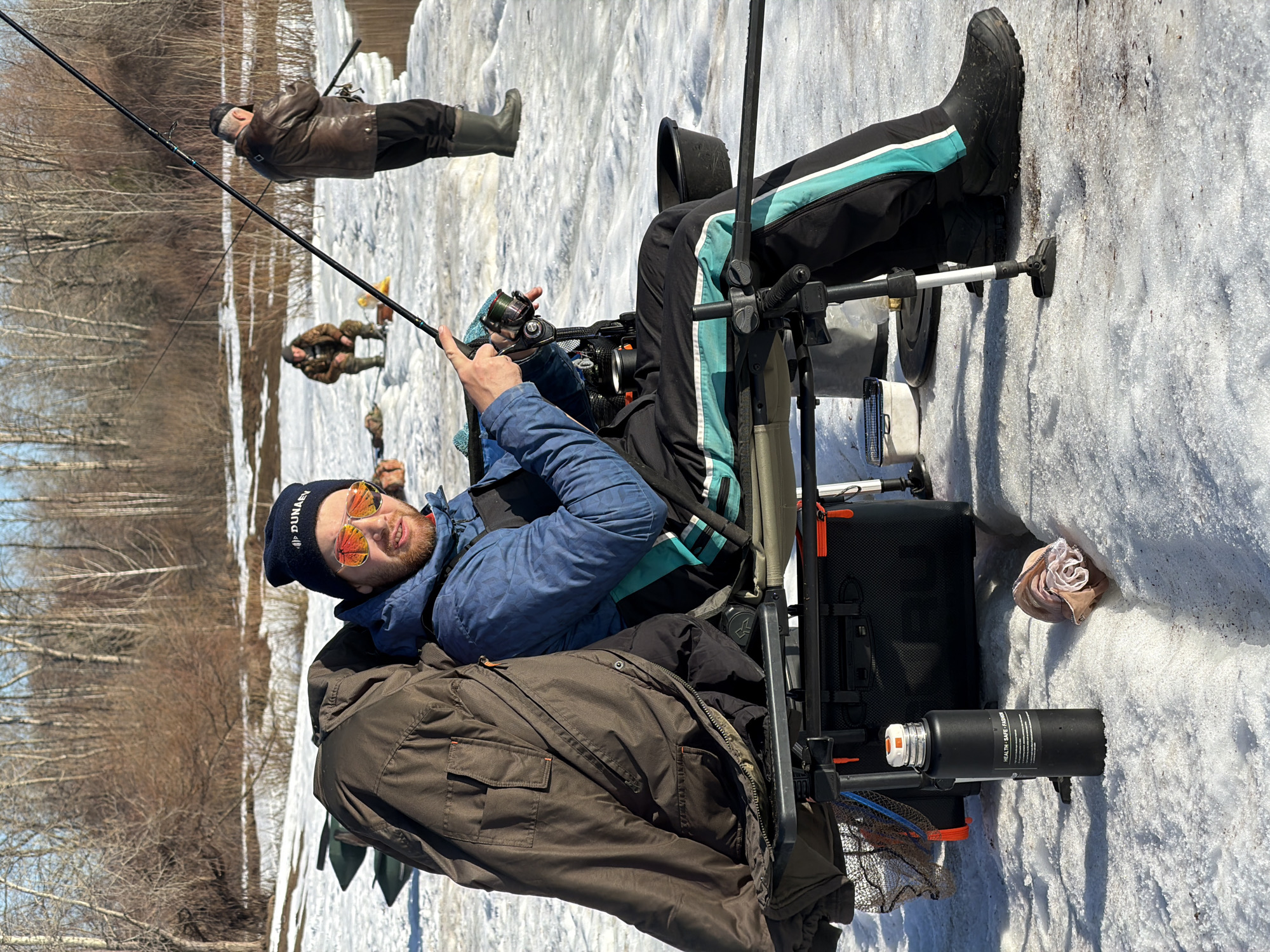 The image shows a winter fishing scene in Noviy, Russia. A man wearing a blue jacket, black hat with the word "Dunay" on it, and orange mirrored sunglasses is seated on a fishing chair with a fishing rod in his hands, apparently ice fishing. He's wearing dark pants with a light blue stripe down the side and black boots. A brown jacket is draped over the chair. Fishing equipment, including a fishing tackle box and a thermos, are next to him. In the background, two other men can be seen fishing on the snowy surface. Trees without leaves are in the background, indicating winter. The ground is covered in snow.