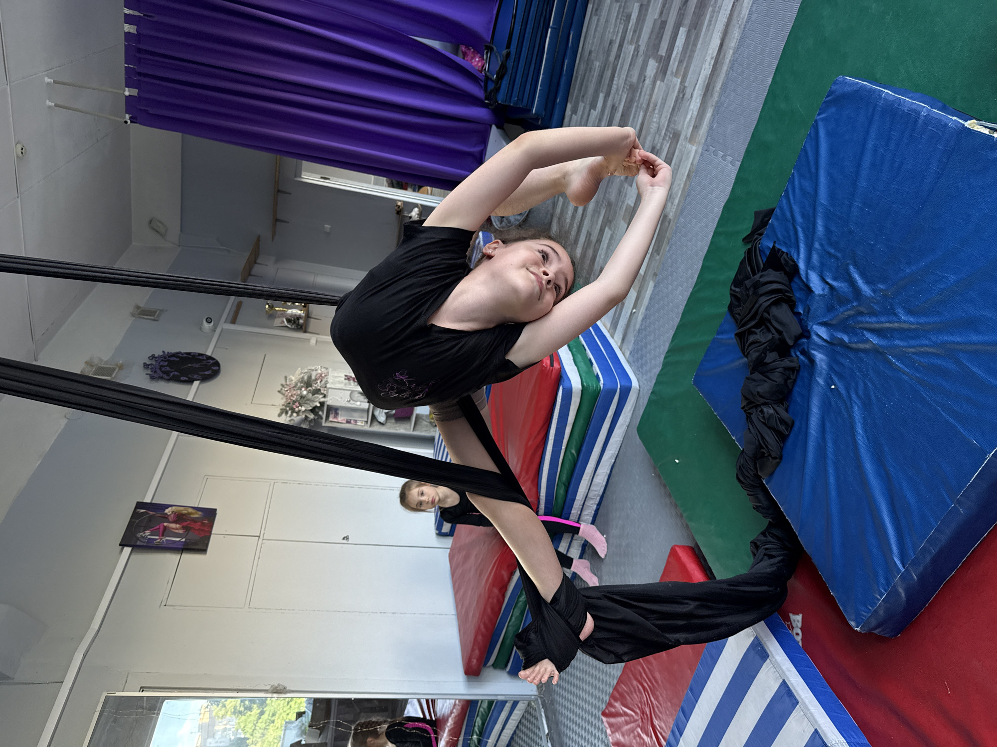 A young girl performs aerial silks in a gym. She is upside down, suspended by black fabric, with her legs bent and arms reaching overhead. Her body is arched in a flexible pose. She is wearing a black t-shirt and black leggings.
In the background, another young girl with blonde hair sits on a red mat. She is wearing a black leotard with pink accents and pink socks.
The gym has a checkered floor and colorful mats, including blue, green, and red. Purple curtains hang on the wall. There is a mirror on the floor reflecting the scene. A small framed picture of a woman in a red outfit is visible on the wall. The room appears to be indoors, likely a dance or gymnastics studio.
The lighting suggests daytime. The overall mood is one of athleticism and performance. No visible text is discernible in the image.