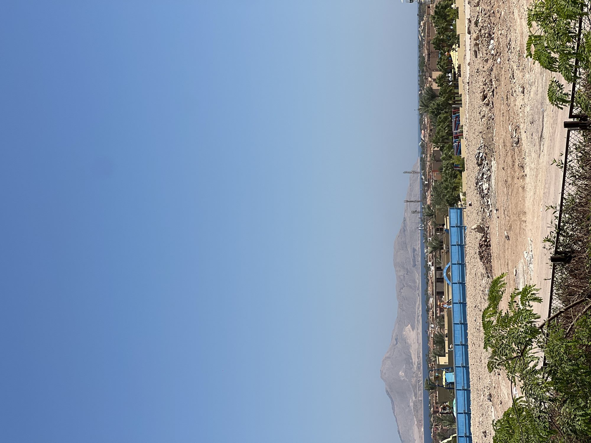 This is a medium-shot, eye-level, outdoor photograph captured on a clear, sunny day. The scene depicts a construction site in the foreground, with piles of dirt, rubble, and construction materials scattered across a barren landscape. A blue barrier fence runs across the middle ground, with construction workers visible in the distance. Beyond the construction site, a resort-like development with buildings and palm trees is visible, situated along the coast of the Red Sea. In the background, a mountain range with rugged, arid terrain rises against the horizon. The sky is a clear, vibrant blue. The location is identified as Unknown, Egypt.