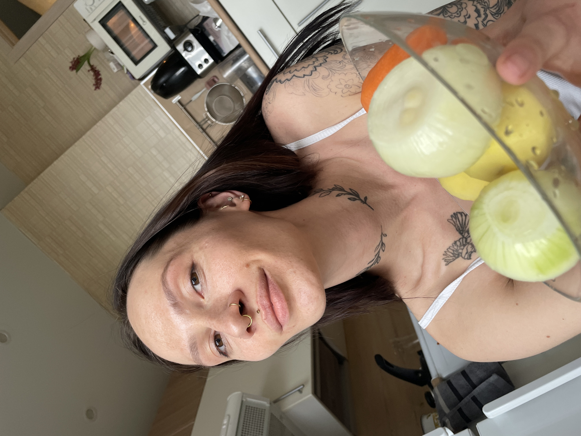 This image depicts a female individual in a domestic kitchen setting, identified as being in Izhevsk, Russia.
The individual is positioned centrally, facing forward and slightly upward, making direct eye contact with the camera. She presents a slight closed-lip smile. Her long, dark hair is parted, with some strands obscuring her right shoulder. She wears a white spaghetti-strap top. A gold-colored U-shaped septum piercing is present. A gold stud is located centrally above the upper lip (medusa piercing). The left ear displays multiple gold-colored piercings, including a small hoop and several studs. Tattoos are visible on the left side of the neck (a delicate leafy branch), the upper left chest (a bird-like figure with wings spread), and the left shoulder/upper arm (a large, intricate floral and abstract design).
Her left hand holds a transparent glass bowl angled towards the viewer. The bowl contains five peeled vegetables: two white onions, two yellow potatoes, and one orange carrot.
The background reveals a well-lit kitchen environment. Features include light-toned kitchen cabinets with metallic handles, a beige tiled backsplash with a subtle grid pattern, and a light-colored countertop. Appliances visible on the counter include a white microwave oven with a dark door on the right, and a black coffee maker or similar small appliance to its left. A stainless steel pot is partially visible behind the individual's left shoulder. A black gooseneck faucet is visible above a sink area to the left. A folded grey towel with two black stripes hangs over a counter edge. A small vase with red foliage is present near the microwave. Two recessed ceiling lights are also visible.