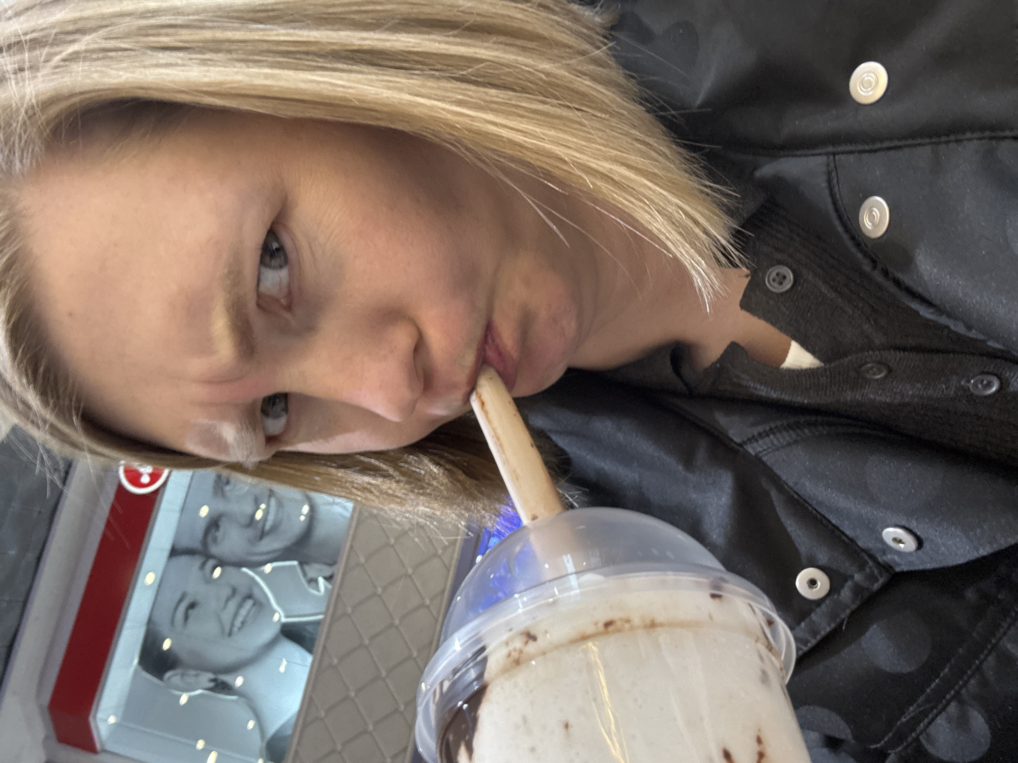 A woman with light blonde hair is drinking a chocolate milkshake through a straw. She is wearing a black jacket with snap closures. In the background, partially visible, is an advertisement or poster showing two smiling people. The location appears to be in Yekaterinburg, Russia, based on the inclusion of a red and white sign for a brand or store prominent in that city.