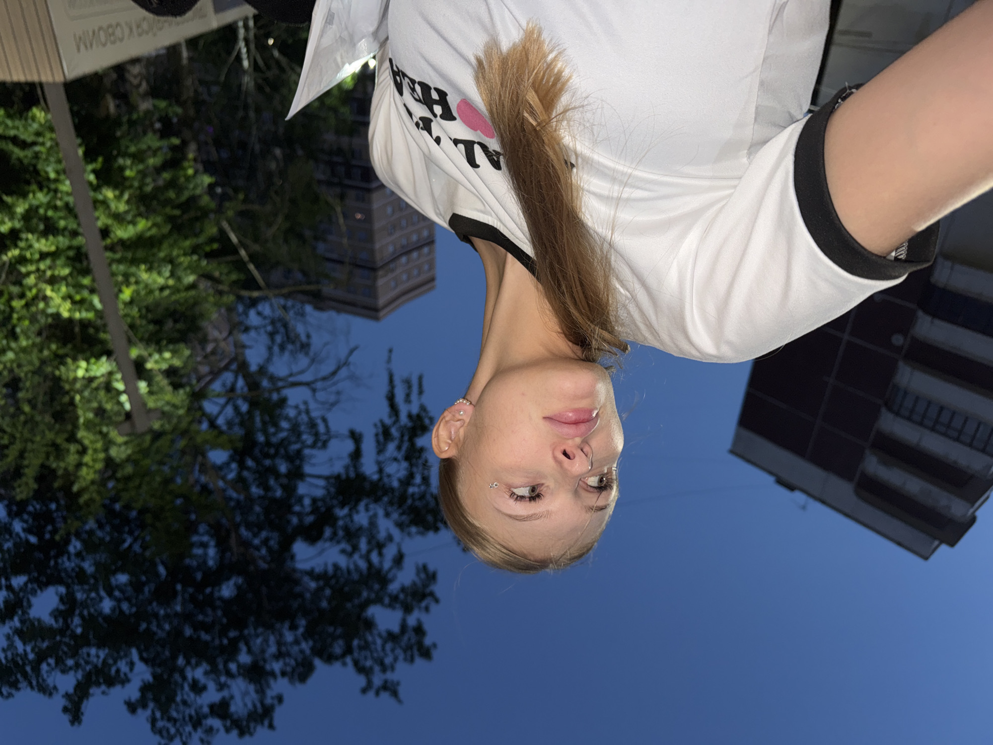 The media file is a selfie taken outdoors at dusk in Perm, Russia. The main subject is a young woman with blonde hair, wearing a white t-shirt with black trim. She has piercings on her ear and face. Her hair is hanging down, partially obscuring her face and the t-shirt. The t-shirt has black text printed on it, along with a pink heart shape.
The background shows a deep blue sky, indicating it is evening. To the left, a large, dark tree with illuminated leaves is visible, suggesting ambient lighting from a streetlamp or building. In the distance, buildings are visible, one appearing to be a multi-story apartment complex. The overall impression is that the photo was taken on a street or in a park within a city. The woman's expression is neutral.