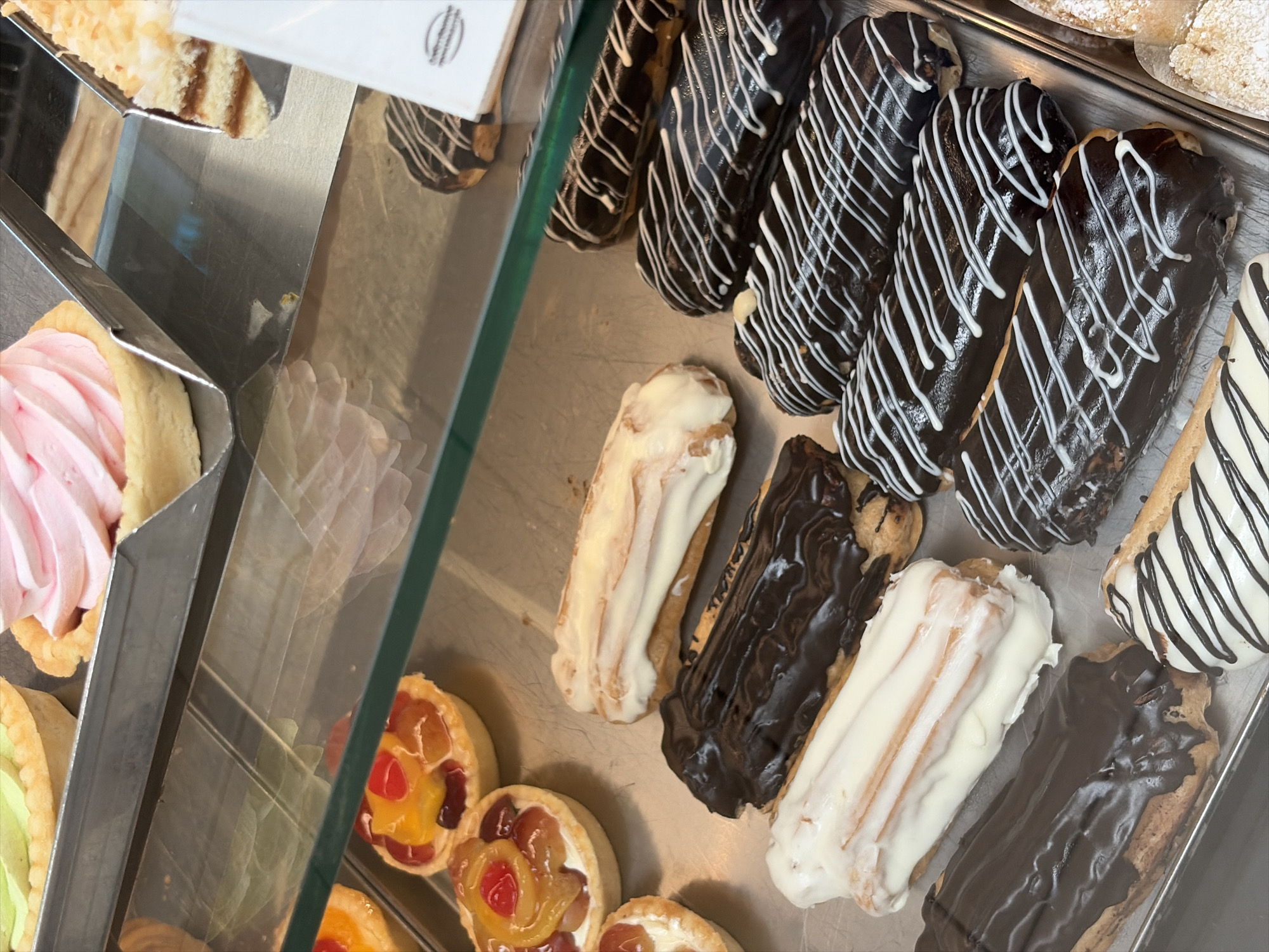 The image displays a close-up view of a bakery display case filled with various pastries. In the foreground, several eclairs are visible, some coated in dark chocolate with white icing drizzle, and others in white icing. Behind them, tarts with fruit toppings are arranged. Further back, a pink swirled pastry and a layered cake with shredded coconut are partially visible. The display case is made of metal and glass, with a green-tinted shelf separating different levels of pastries. The lighting suggests an indoor setting, likely a bakery. No people are visible in the image. The scene depicts a selection of desserts ready for purchase. This bakery is located in Balashikha, Russia.