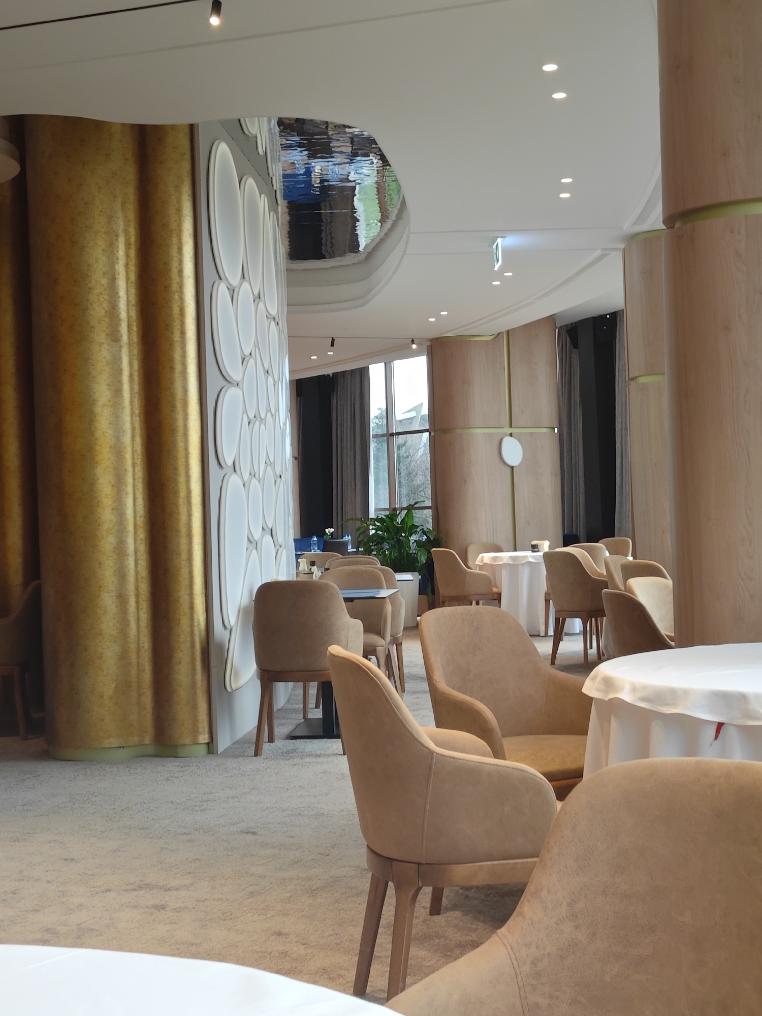 The image depicts the interior of a restaurant. The location is likely in Varna, Bulgaria based on the known association of this style with restaurants in that city. The space is furnished with round tables draped in white tablecloths, surrounded by upholstered armchairs with wooden legs. Walls are adorned with textured panels in gold and white. There is a shiny, reflective ceiling feature. A view through a window reveals greenery outside. No people are visible. The lighting is bright and even.