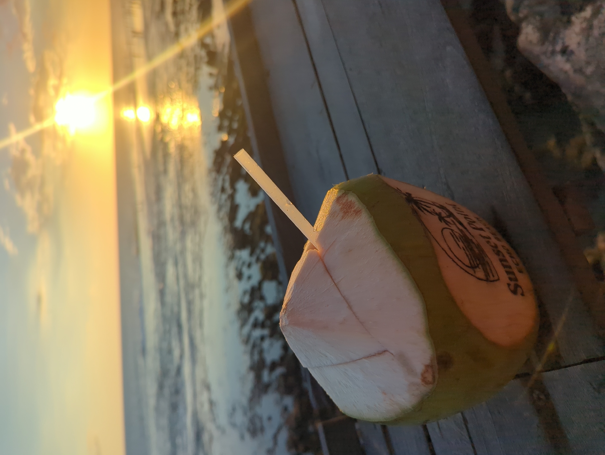 The image shows a half-cut coconut with a straw, resting on a wooden surface. In the background, there is a beach scene with the sun setting, reflecting on the water. The name "Sunset Hotel" is visible on the side of the coconut. The setting is outdoors, and the general location appears to be a coastal area in North Kuta, Indonesia, as suggested by the name on the coconut.