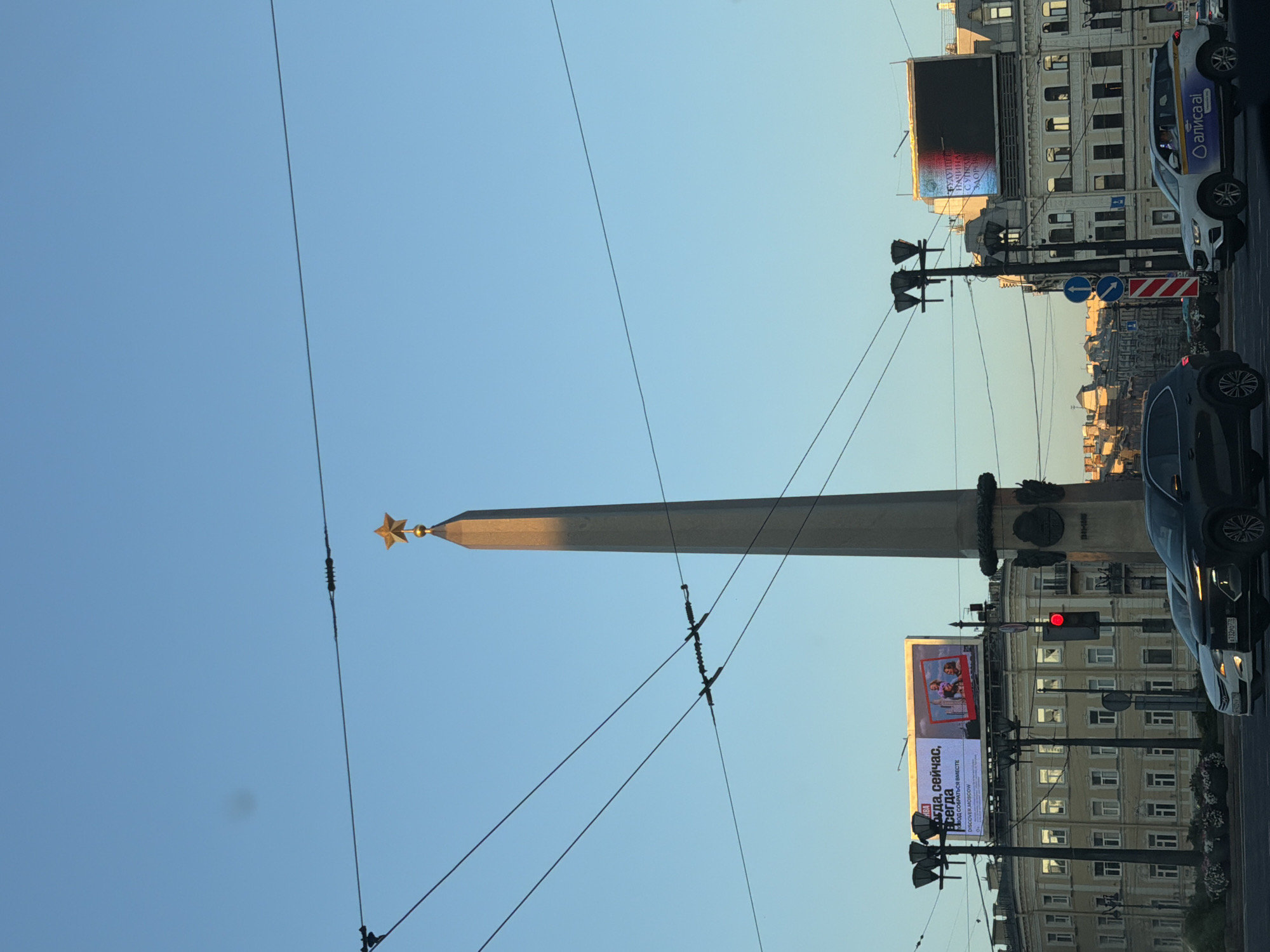 The image depicts a view of the city of Saint Petersburg, Russia, specifically featuring the obelisk at Victory Square (Ploshchad Pobedy). The obelisk, topped with a golden star, stands prominently against a clear blue sky. Several overhead wires, likely for public transportation, crisscross the frame. In the foreground, a street scene unfolds with cars, including a white taxi with "АЛИСА ai" branding, navigating the road. Traffic lights are visible, with one showing red. Buildings line the street, one featuring a large digital billboard displaying text in Russian and an image. The overall lighting suggests it is either early morning or late afternoon, casting a warm glow on the scene.
