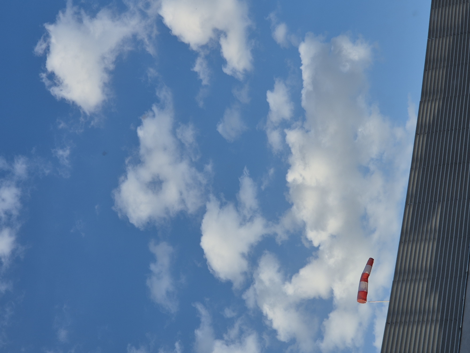 A vibrant blue sky, partially filled with scattered white cumulus clouds, dominates the upper two-thirds of the vertical frame. The clouds are varied in size, with some appearing brighter and others exhibiting subtle grey shading. The lower portion of the image reveals the top edge of a grey, horizontally corrugated building or wall. Mounted on a thin, light-colored pole, and extending above this structure into the sky, is a red and white striped windsock. The windsock is fully inflated and oriented horizontally, indicating the presence and direction of wind. The scene is illuminated by natural daylight in the city of Unknown, Unknown.