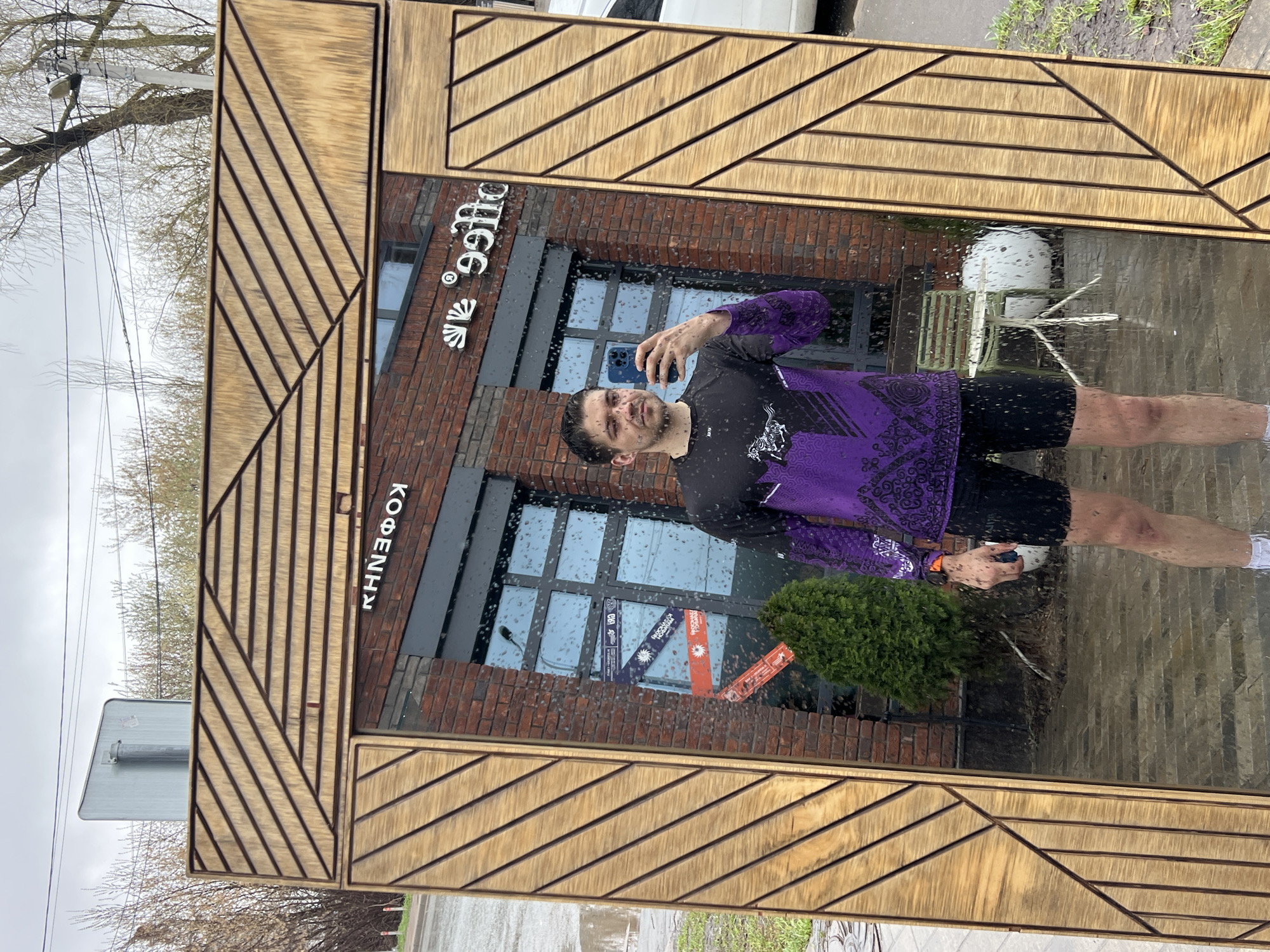 A man is taking a selfie in a mirror outside a coffee shop in Kazan, Russia. The coffee shop has a brick facade with the word "Кофейня" (Coffee Shop) vertically on the left and "coffee" horizontally on the top. The window displays promotional materials. The man is wearing a black and purple athletic shirt and black shorts, holding a smartphone. A small tree is positioned outside the window. The weather is overcast and it appears to be raining.