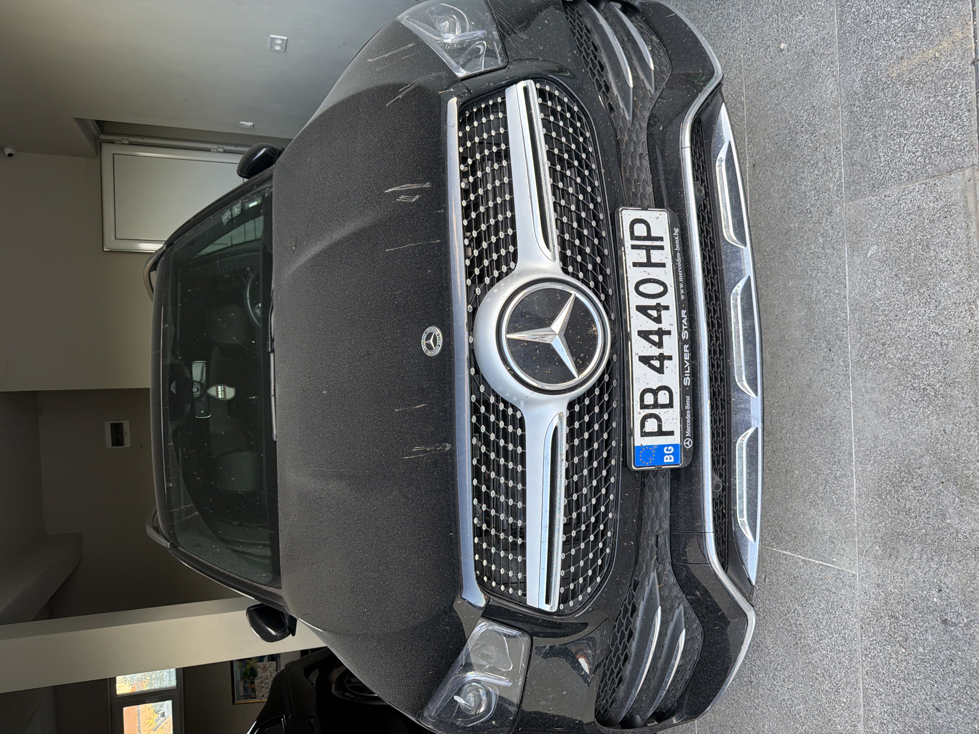 A close-up shot captures the front of a black Mercedes-Benz SUV. The car's grill, emblazoned with the Mercedes logo, dominates the frame. The license plate reads "PB 4440 HP" and includes "BG" for Bulgaria. The bumper and headlights are visible, appearing dusty. The car is parked on a grey stone floor, seemingly inside a garage or a covered area. While the precise location in Asenovgrad, Bulgaria cannot be determined, the Bulgarian license plate suggests it is located in Bulgaria.
