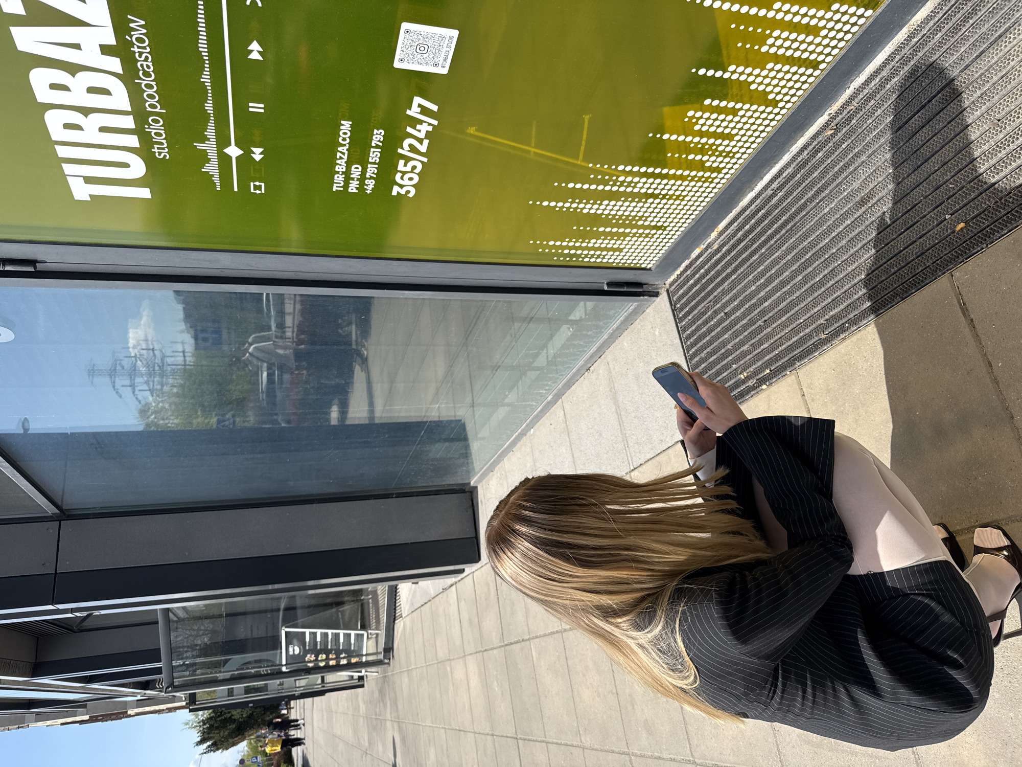 A woman with long blonde hair is seated on the sidewalk in front of a building with a large window displaying an advertisement for "TURBAZ" studio podcasts. She is wearing a black pinstripe suit and holding a mobile phone in her hands. The advertisement on the window includes a play/pause symbol, a QR code, and the text "TUR-BAZA.COM," "PN-ND," "+48 791 551 793," and "365/24/7." The window reflects the surrounding urban environment. The location is identifiable as Warsaw, Poland due to the "+48" phone code.