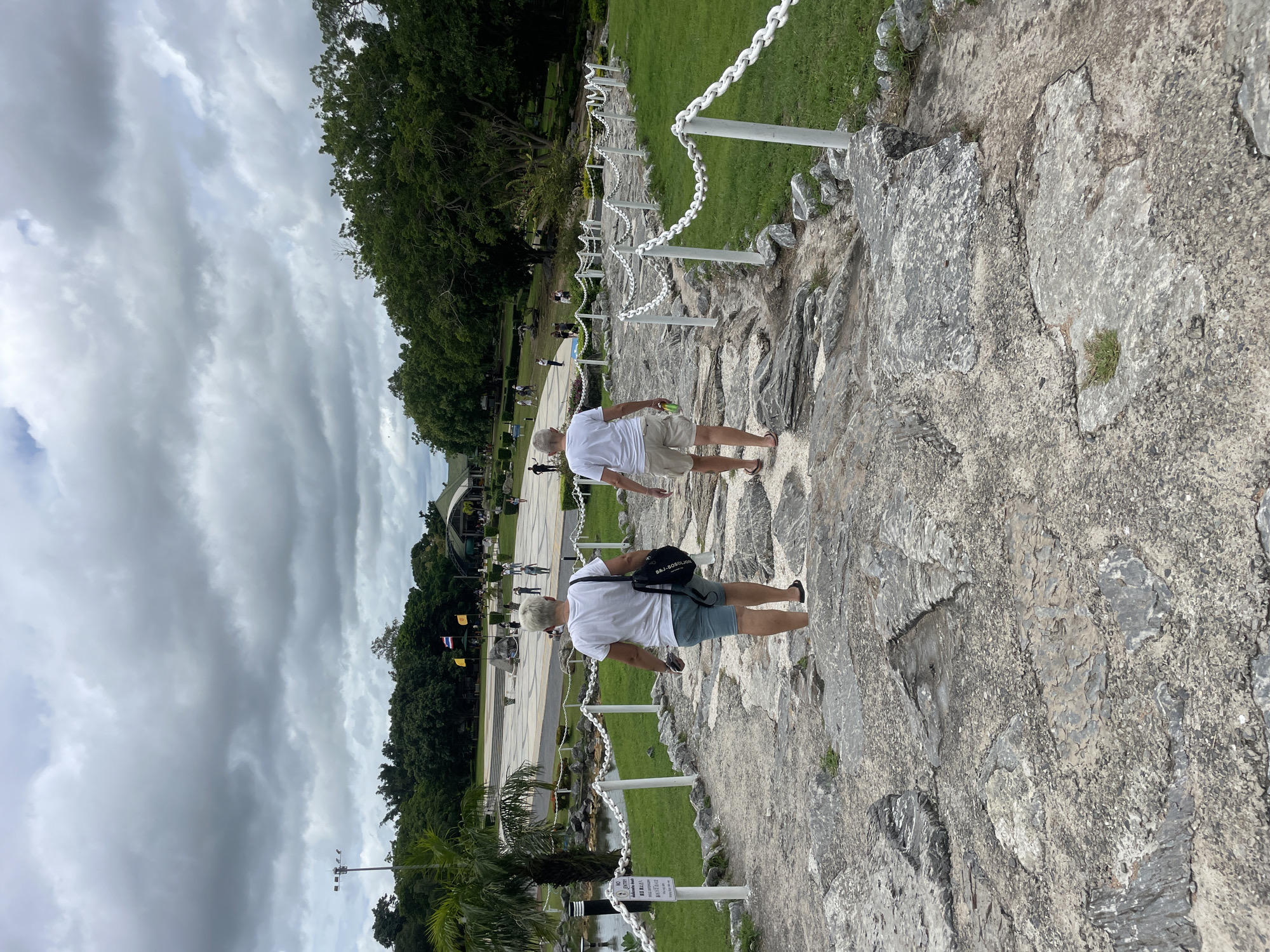 Two individuals, appearing to be adults, are walking up a rough stone pathway bordered by a white chain fence. Both are wearing white t-shirts and casual shorts. The person in the foreground is wearing light blue shorts and carrying a black bag over their shoulder, while the person behind them is wearing tan shorts and holding a green object in their right hand. The pathway leads uphill towards a paved area with several people in the distance and a large, open-sided building with a green roof. The environment is a park-like setting with lush green grass, trees, and palm trees. The sky is overcast with dense grey clouds. A sign with text is visible near the bottom left of the pathway. The location is identified as Na Chom Thian, Thailand, suggesting this is a public park or tourist attraction.