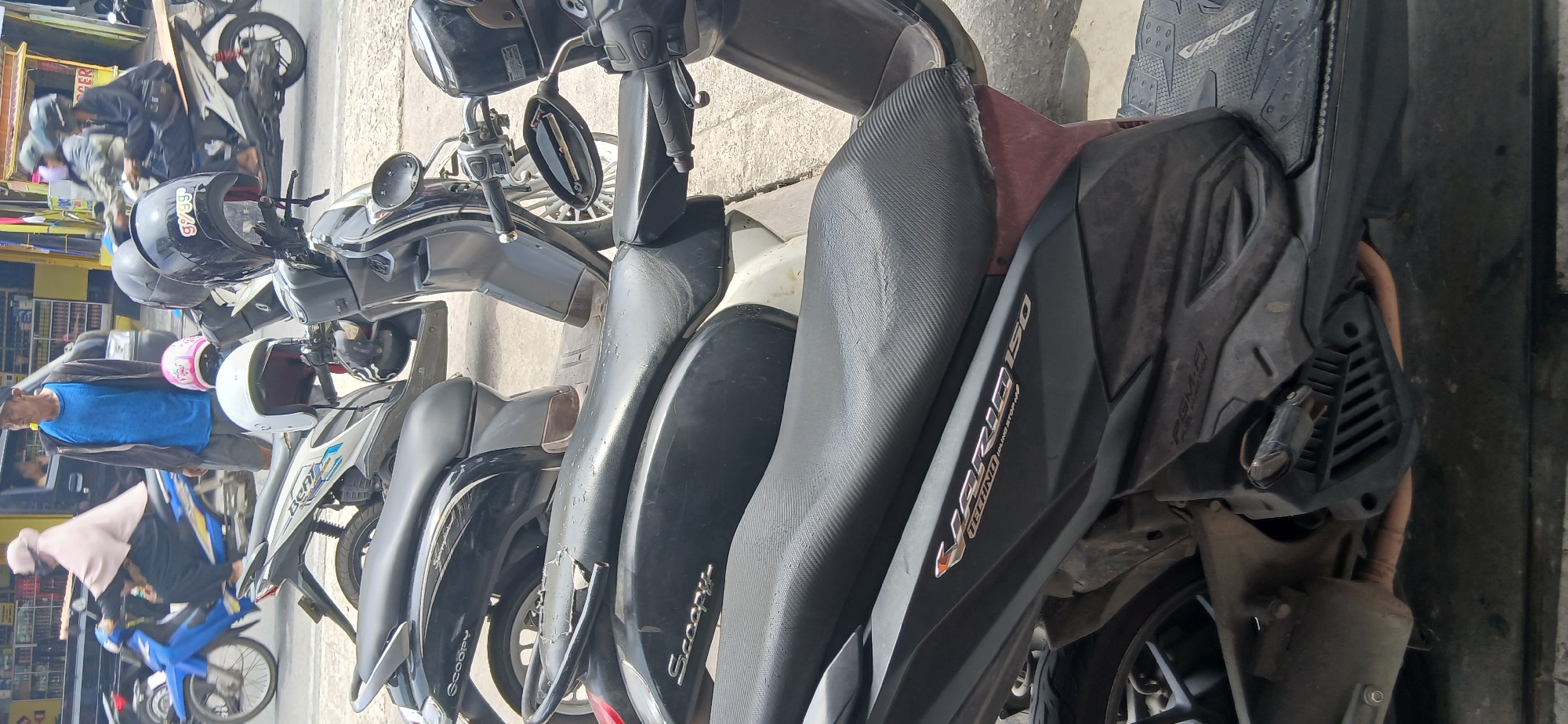 The image depicts a street scene in Samarinda, Indonesia, with several motorcycles parked on a paved surface. In the foreground, a dark-colored scooter with a black seat is prominently featured, with the "VARIO 150" logo visible on its side. To the left, another dark scooter with the "Scoopy" logo is partially visible. Behind these, several other motorcycles are parked, including a white and blue scooter.
A man wearing a blue shirt and a dark cap stands near the parked motorcycles, looking towards the right. In the background, a woman in a pink headscarf is riding a blue motorcycle, and other vehicles and buildings line the street, suggesting an urban environment. The overall impression is a typical street view in a Southeast Asian city, with motorbikes being a common mode of transportation.