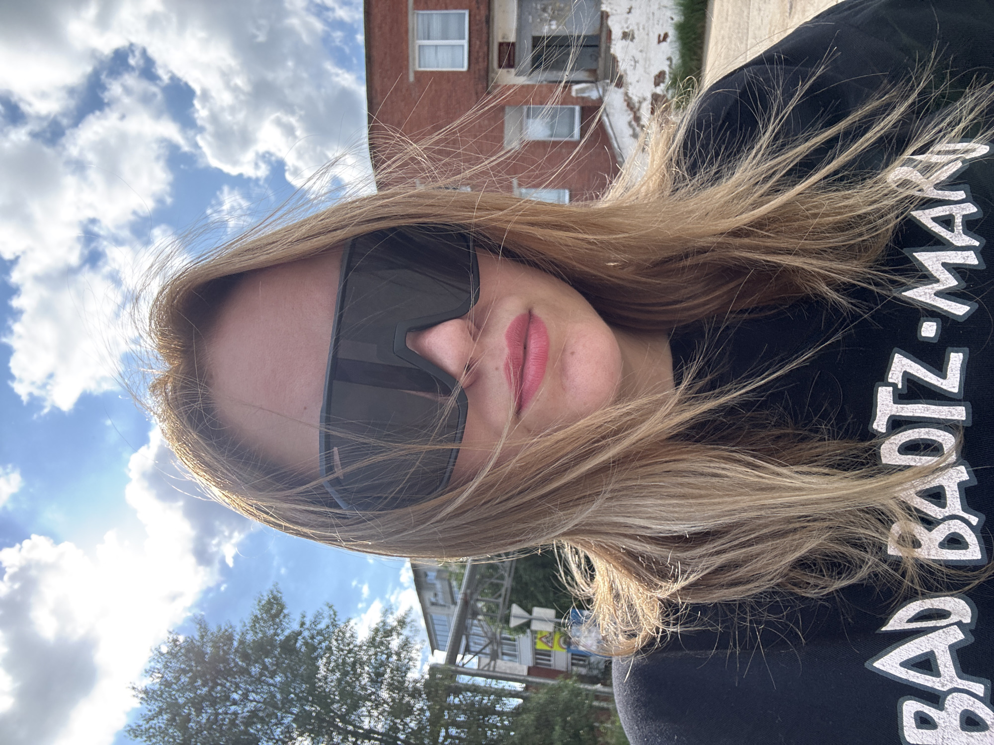 A woman is taking a selfie outdoors, with a blue sky and clouds in the background. She is wearing large, dark sunglasses and a black t-shirt with white lettering that reads "BAD BADTZ-MARU". Her blonde hair is windblown. Behind her, to the left, is a green tree and a street sign. To the right, a red brick building with several windows is visible. The setting appears to be a street in Noviy, Russia, possibly near residential buildings.