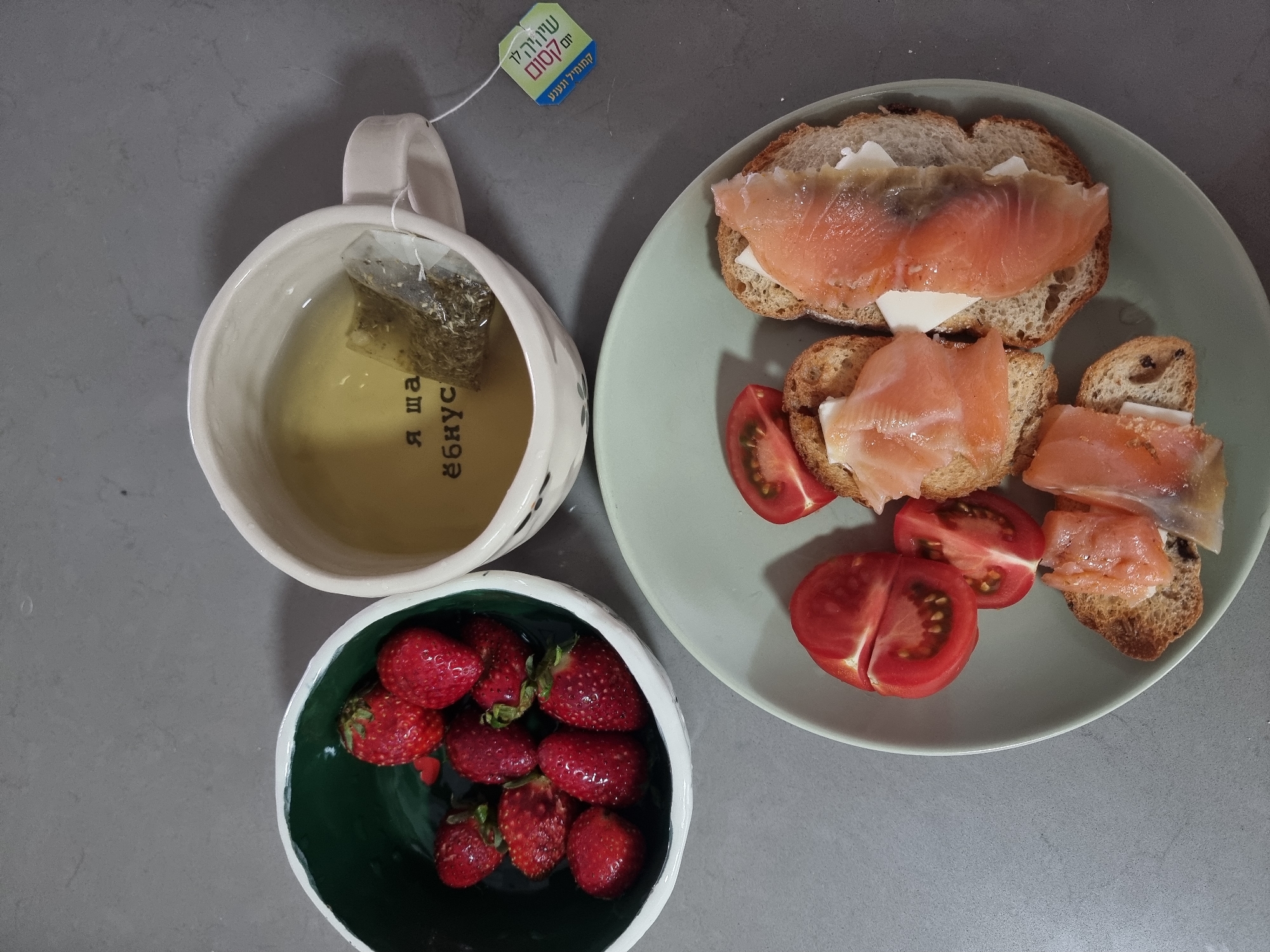 The image shows a still life of a meal consisting of a cup of tea, a bowl of strawberries, and a plate of open-faced sandwiches with salmon and tomatoes. A tea bag with Hebrew writing is visible near the cup. The photo was taken in the city of Ramat Gan, Israel.