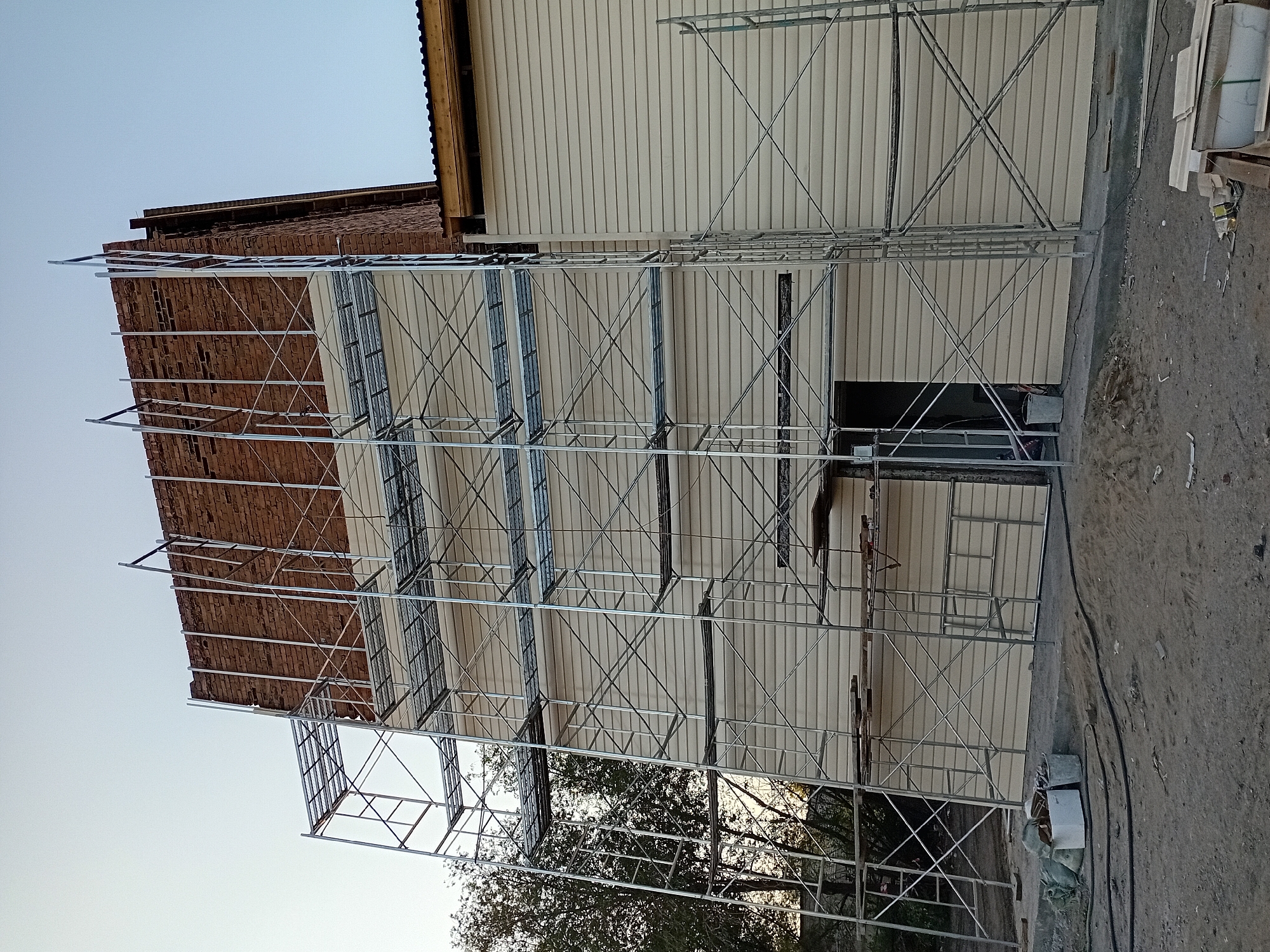 The image shows the exterior of a building under construction or renovation in Semey, Kazakhstan. A large scaffolding structure is erected against the side of the building, which is clad in light-colored siding. A portion of the building's upper section is constructed with red brick. The ground in front of the building is a dirt and gravel mix, with some construction debris and materials visible, including bags of sand, buckets, and lumber. Trees and vegetation are visible to the left of the building, suggesting an outdoor setting. The sky is clear and appears to be either early morning or late afternoon.