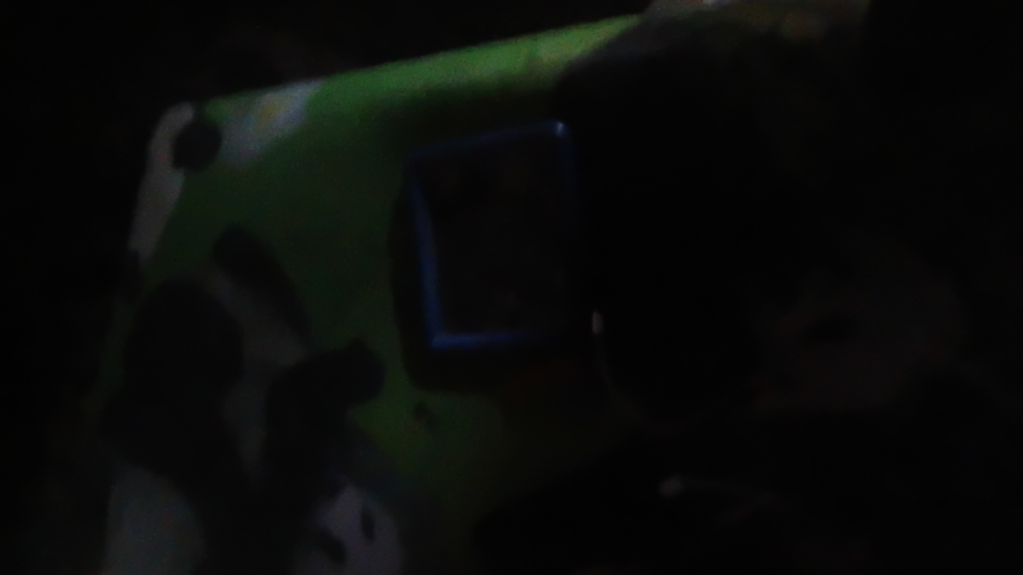 The image depicts a dark, blurry scene with a hint of a green surface patterned with irregular dark shapes. In the center, a rectangular object with a blue border is visible, possibly a screen or a device. The overall lighting is very low, making it difficult to discern specific details. There is no discernible location information within the image that would suggest it is from Prabumulih, Indonesia.