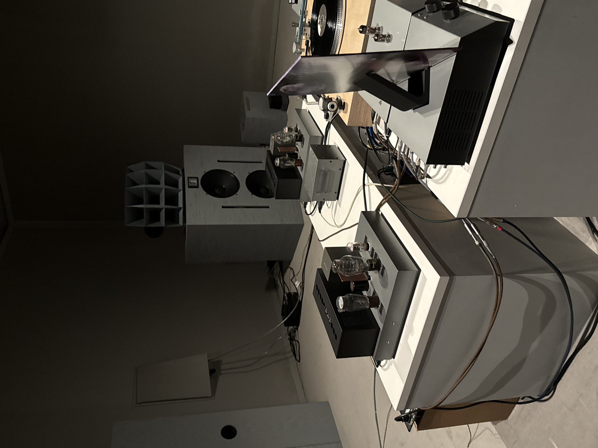 This media file presents a high-fidelity audio system meticulously arranged in a dimly lit indoor space, likely a dedicated listening room or studio. The setting features light grey walls and multi-tiered white shelving that hosts the audio components.
The central subjects are a collection of professional-grade audio equipment. Two prominent, light grey loudspeakers are visible, one in the mid-ground and another partially obscured. Each speaker sports two large, dark-coned drivers and a rectangular detail near the top. On the floor beside the main speaker, a dark grey acoustic diffuser with a geometric, stepped pattern is strategically placed for sound optimization.
Further into the background, a turntable with a black vinyl record rests on a wooden platform, integrated into a larger grey and white audio rack system. Below this, and on an adjacent shelf, are several other critical audio components. Two tube amplifiers or pre-amplifiers stand out, their exposed vacuum tubes glowing softly, signaling their operational status. These are accompanied by other black and grey units, including what appears to be a power supply or additional amplification, and various interconnected devices.
A dense network of cables, including power cords and audio interconnects, weaves through the setup, linking all the components. The aesthetic is clean and modern, combining light grey, white, and black elements with the warm wood tone of the turntable base. The dim lighting conditions create an intimate atmosphere, drawing attention to the glowing tubes and the intricate arrangement of the system. While the location context is London, United Kingdom, no specific visual cues within the image confirm this. There is no visible text on any of the equipment or surfaces. The overall scene suggests a sophisticated environment tailored for an immersive music listening experience.