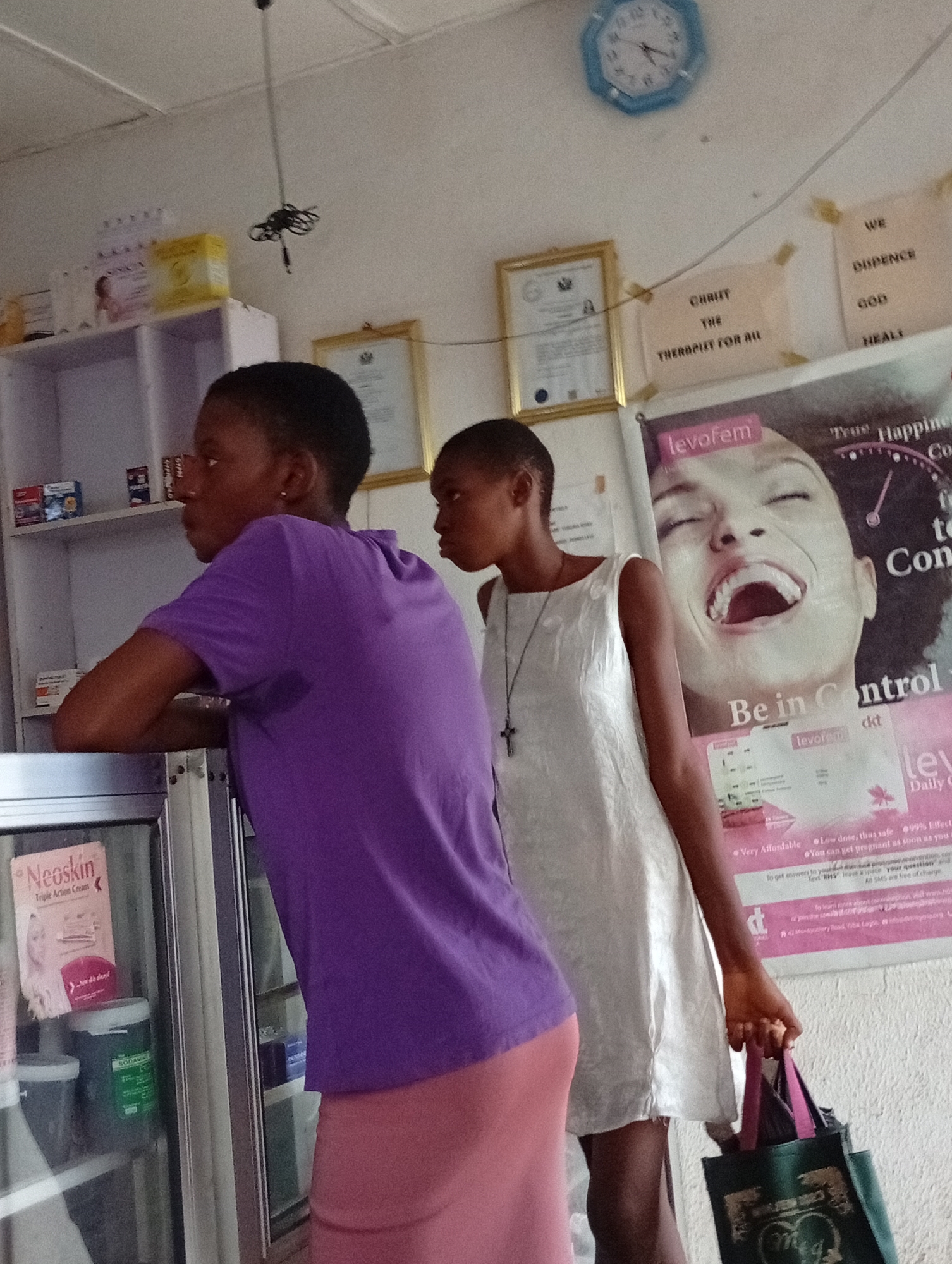 In an indoor setting, likely a small shop or pharmacy in Adankolo, Nigeria, two young women are captured in a candid moment. The woman in the foreground, wearing a purple t-shirt and a pink skirt, leans against a display counter, facing left in profile. Her arm rests on the counter's edge. Behind her, another young woman in a white sleeveless dress, adorned with a cross pendant, stands holding a dark green tote bag in her right hand. Both women have short, dark hair.
The setting includes shelves stocked with various products to the left of the frame. The display counter in the immediate foreground shows a "Neoskin Triple Action Cream" product and a green jar labeled "SODAWN." The background wall is adorned with several items: a light blue wall clock, two framed documents (possibly certificates), and multiple posters.
Notable text includes religious messages on beige paper, reading "CHRUT THE THERBOUT FOR AU" (likely "CHRIST THE THERAPIST FOR ALL") and "WE DUPENCE GOD HEALI" (likely "WE DEPEND ON GOD HEALS"). A large, prominent pink and black poster for "levofem" features a smiling woman's face and text such as "True Happine," "Be in Control," "Very Affordable," "Low dose, thus safe," "99% Effect," "You can get pregnant as soon as you," and details about texting for questions and free SMS. The clock on the wall indicates a time close to 2:45. The overall atmosphere suggests a casual, local business environment.