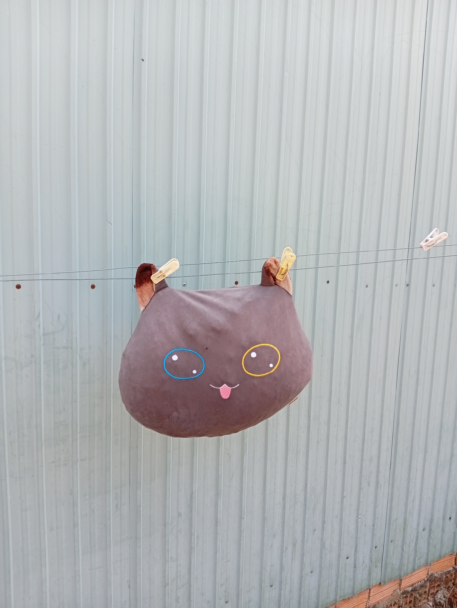 A medium shot depicts a gray plush cat-shaped pillow suspended from a clothesline by two clothespins against a background of vertically-paneled, light blue metal siding. The pillow has two small brown ears held by the clothespins. The cat's face is embroidered with a blue circle around its left eye and a yellow circle around the right eye. A small pink tongue is sticking out from the mouth. The clothesline is strung across the paneling, and a third clothespin hangs alone to the right of the pillow. The scene is at Phú Bình, Vietnam, evident due to 'MADE IN VIETNAM' written on the cushion. The bottom of the siding shows a row of orange bricks.