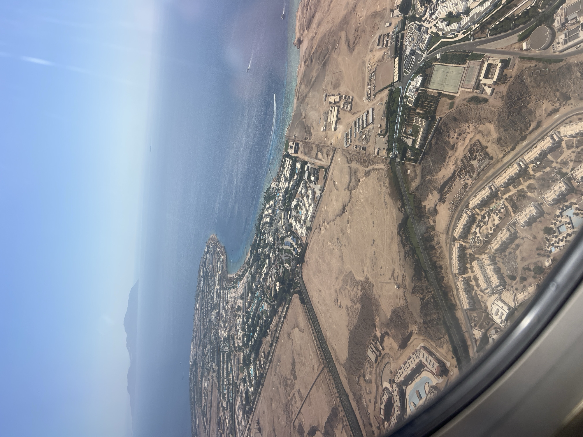 This aerial photograph captures a section of the coastal city of Sharm El Sheikh, Egypt, viewed from an airplane window. The scene is dominated by the contrasting landscapes of the arid desert and the vibrant blue of the Red Sea. Numerous resort complexes with white buildings and swimming pools are scattered across the desert terrain, many situated along the coastline. The resort architecture features multiple interconnected buildings, often arranged around courtyards or open spaces. Roads and pathways weave through the complexes and connect them to the main thoroughfares. The Red Sea is a prominent feature, its clear waters revealing shallow reef areas near the shore. Several boats, including a speed boat leaving a wake, are visible on the water. In the distance, a mountainous island or landmass is silhouetted against the horizon. The photograph is taken during daylight hours, with clear skies and bright sunlight casting shadows. The perspective suggests the aircraft is in the process of descending or ascending, given the angle and the inclusion of the airplane window frame in the foreground.