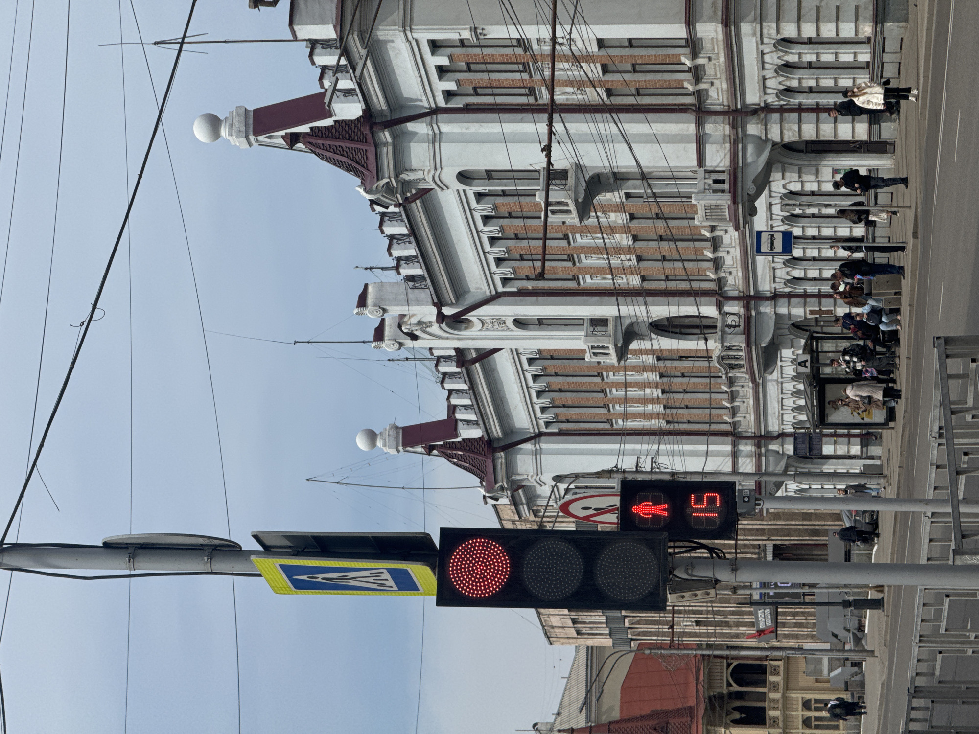 The image depicts an urban street scene in Krasnoyarsk, Russia, under a clear, bright sky. The photo is taken from a street-level perspective, focusing on an intersection with a large, ornate building on the right.
In the foreground, a utility pole holds traffic signals. A main traffic light displays a solid red circular light, while a pedestrian signal to its right shows a red standing human figure and a numerical countdown of "15" seconds. Attached to the pole is a yellow and blue sign indicating a pedestrian crossing. Overhead, numerous power and communication lines crisscross the sky, suggesting the presence of public transport like trolleybuses or trams.
The prominent building on the right is a multi-story structure characterized by intricate classical architecture. It features a light-colored facade with decorative elements such as cornices, pilasters, and arched window frames, interspersed with brickwork. The roofline includes decorative white spherical finials and dark-colored, possibly red, accents.
On the sidewalk in front of this building, several people are visible. Some are gathered near a bus stop, marked by a blue square sign with a white bus icon. Others are walking or standing, all dressed in coats and jackets, indicating cooler weather. The street and a portion of the sidewalk are visible in the lower right.
The scene captures a typical daytime urban moment, with pedestrians waiting at a crossing and bus stop against the backdrop of historic city architecture.