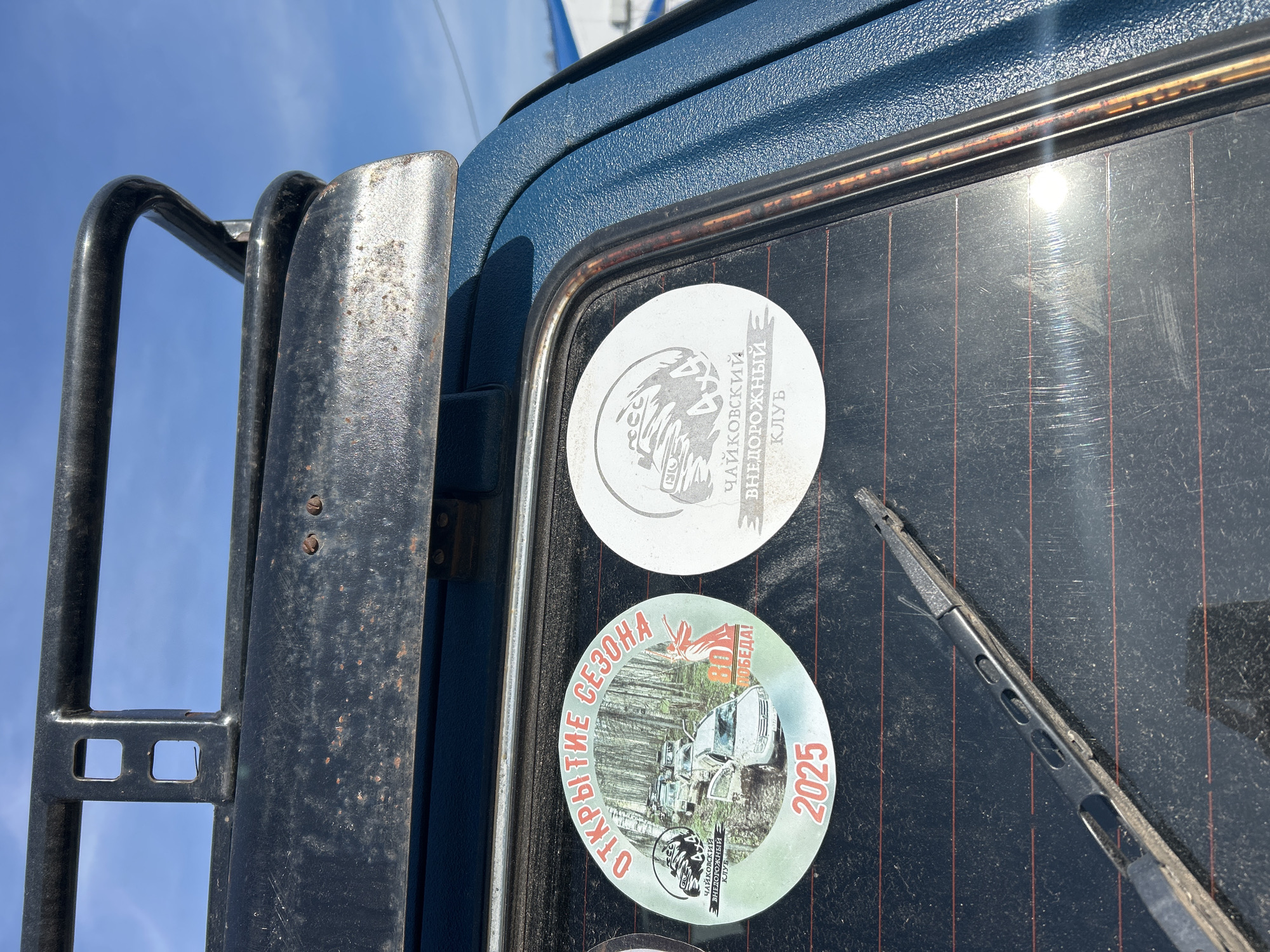 The image shows a close-up of the rear of a vehicle, likely an off-road vehicle based on the visible roll cage and stickers. The vehicle is dark blue and appears textured. Two round stickers are affixed to the rear window. The top sticker features a stylized drawing of a bear and text in Russian. The bottom sticker depicts a line of off-road vehicles in a wooded area and the year "2025". The text "ОТКРЫТИЕ СЕЗОНА" (Opening of the Season) is visible in Russian around the perimeter. The rear window is tinted and has a rear window wiper. The image was taken outdoors, as evidenced by the blue sky. No people are visible. There is no specific information in the photo to confirm its location in the city of Ural'skoe, Russia.