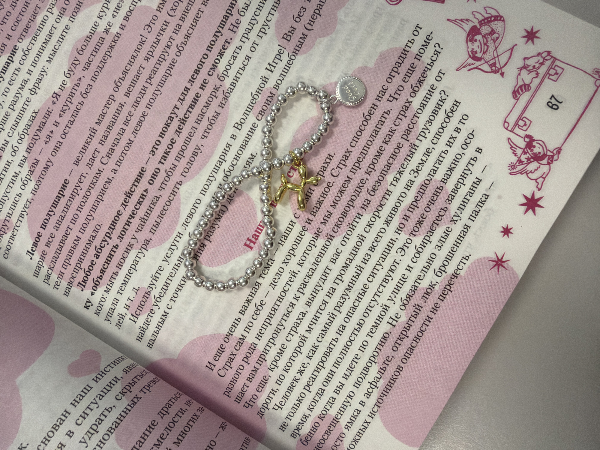This image shows a close-up view of an open book with pink heart-shaped illustrations on the page. The book appears to be a publication with printed text in Russian. A silver bracelet with a circular charm that says "Good Luck" is draped across the open pages. The text discusses various psychological concepts and behaviors, including "left-wing" and "right-wing" behavior, fear, and instinct. There are also small illustrations of cherubic figures and a suitcase with the number 67 on it. The page is white with pink accents. There is no visible indication of the city of Homyel, Belarus, in the image.