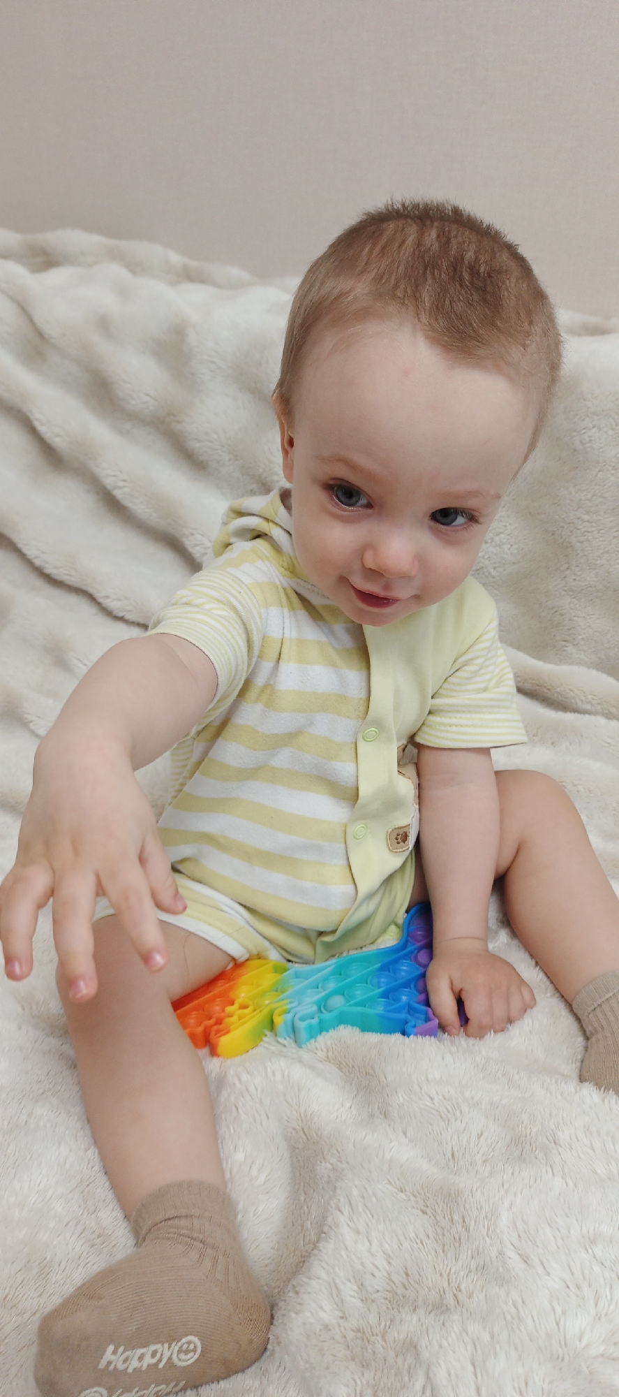 A young child, likely a toddler, with light brown hair and blue eyes, is seated on a fluffy, light-colored (beige or cream) blanket or rug. The child is wearing a light yellow and white horizontally striped short-sleeved onesie or romper, featuring snap buttons down the front and a small brown patch with an animal design, possibly a bear, on the chest. Light brown socks are on the child's feet; the word "Happy" accompanied by a smiley face logo is visible on the sole of the left sock. A vibrant, rainbow-colored "pop-it" fidget toy is positioned between the child's legs, and one of the child's hands is reaching towards it, suggesting engagement with the toy. The child looks directly at the camera with a gentle, curious expression and a faint smile. The background is a plain, light-colored wall, indicating an indoor setting with soft, even lighting.