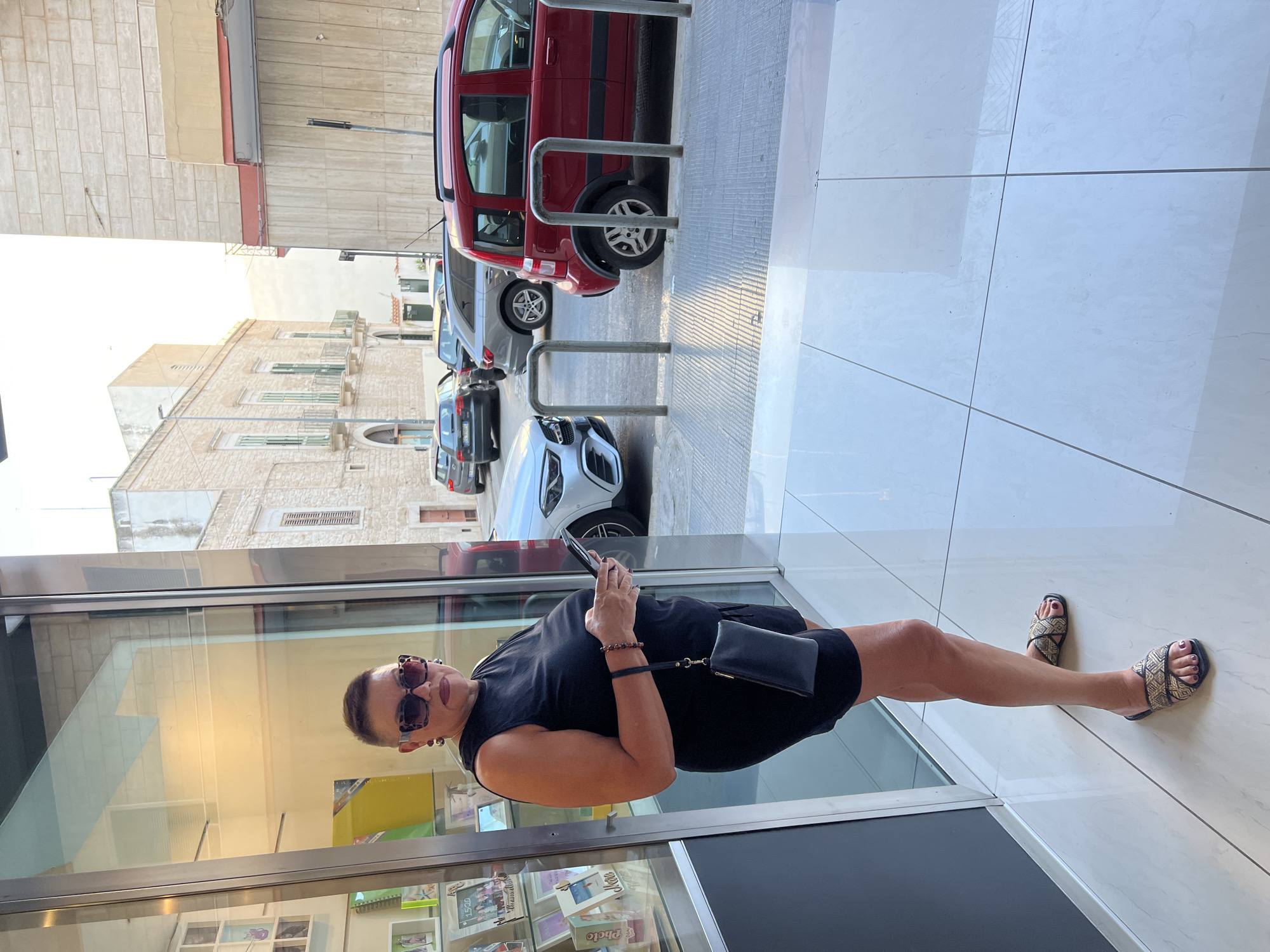 A woman stands in front of a glass-fronted building, holding a smartphone. She is wearing a black dress, sunglasses, and sandals. A black clutch bag is attached to her wrist by a strap. The building's interior is visible through the glass, displaying shelves with books and other items. The exterior shows a tiled sidewalk and a street with parked cars, including a red SUV and a white car. The scene appears to be set during the daytime, with natural light illuminating the area. The architecture of the buildings in the background suggests an urban European setting, consistent with the provided location context of Bitonto, Italy.