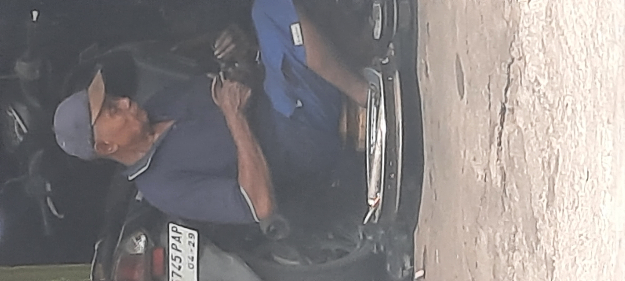 An elderly Indonesian man wearing a dark blue polo shirt, blue shorts, and a blue baseball cap with a brown brim is seated on a motorcycle in Selopuro, Indonesia. He appears to be holding something small in his hands. The motorcycle has a license plate that reads "5745 PAP 04-29". The man is positioned in what appears to be a workshop or a similar environment, with other motorcycles and equipment visible in the background. The ground in front of the motorcycle is paved with concrete.