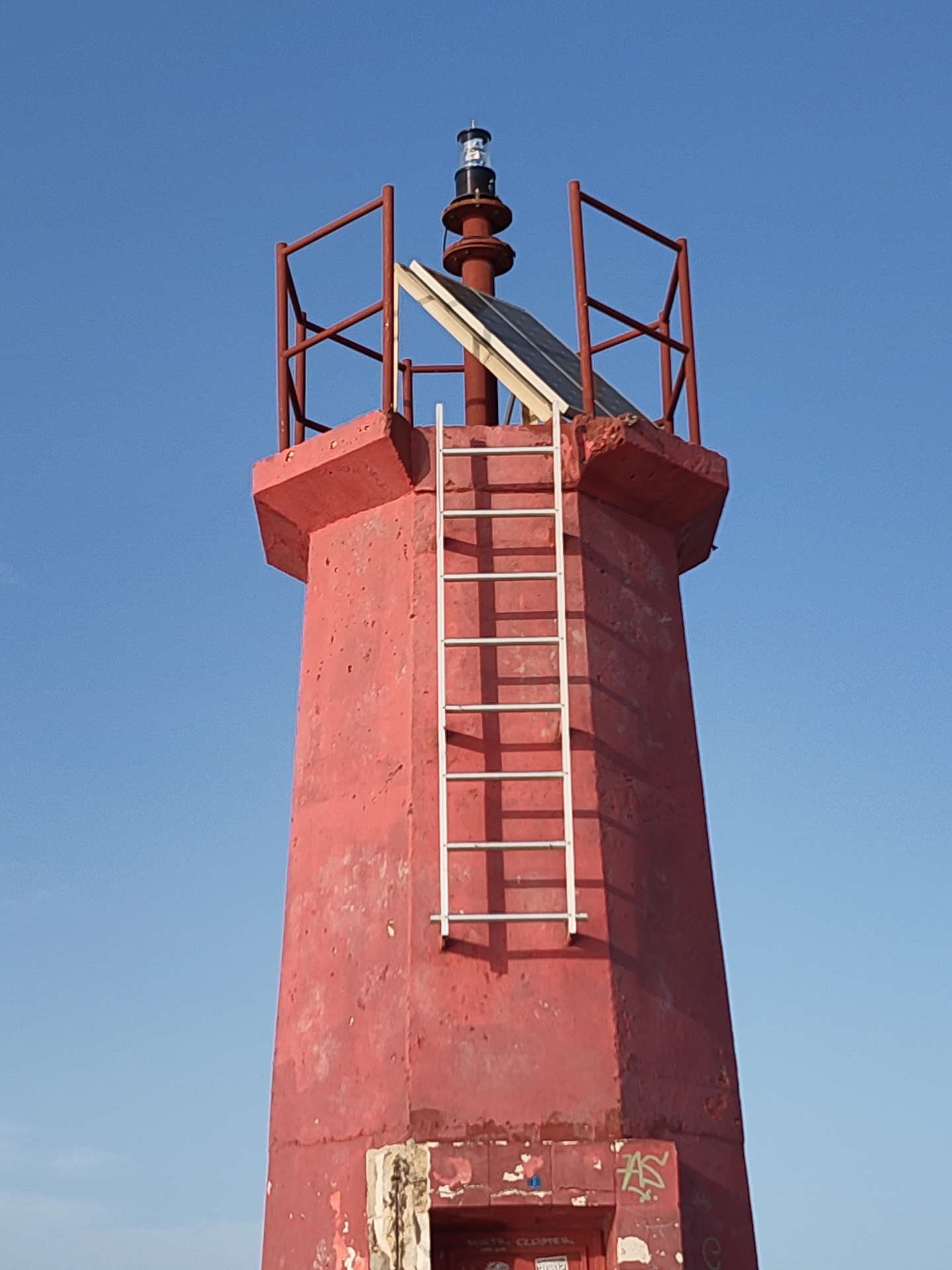 The image shows a red lighthouse tower in Dénia, Spain. The tower is constructed of concrete and has a pyramidal shape with a wider base tapering upwards. A white metal ladder is affixed to one side of the tower, extending from near the base to the top. At the top of the tower, there is a red metal railing and a solar panel positioned near the top. On the very top of the structure, there is a black and clear glass light fixture. The base of the tower has weathered and damaged areas. Some graffiti is visible near the bottom of the tower. The background is a clear, bright blue sky. There are no people visible in the image.