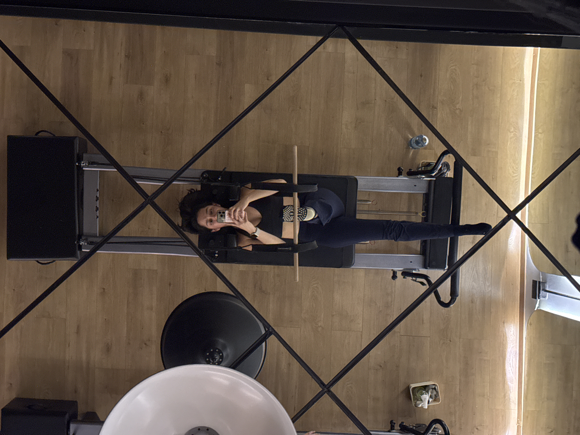 A woman lies on a Pilates reformer machine, taking a selfie with her phone. She is wearing a dark t-shirt and leggings, and has dark hair. The Pilates reformer is set against a light wood floor. A portion of a lighting setup is visible in the lower left, and the scene is framed by a black metal grid, likely the structural element of a studio or gym. The location is likely indoors, possibly in a Pilates studio in Puebla, Mexico, though no specific location indicators are visible in the image.