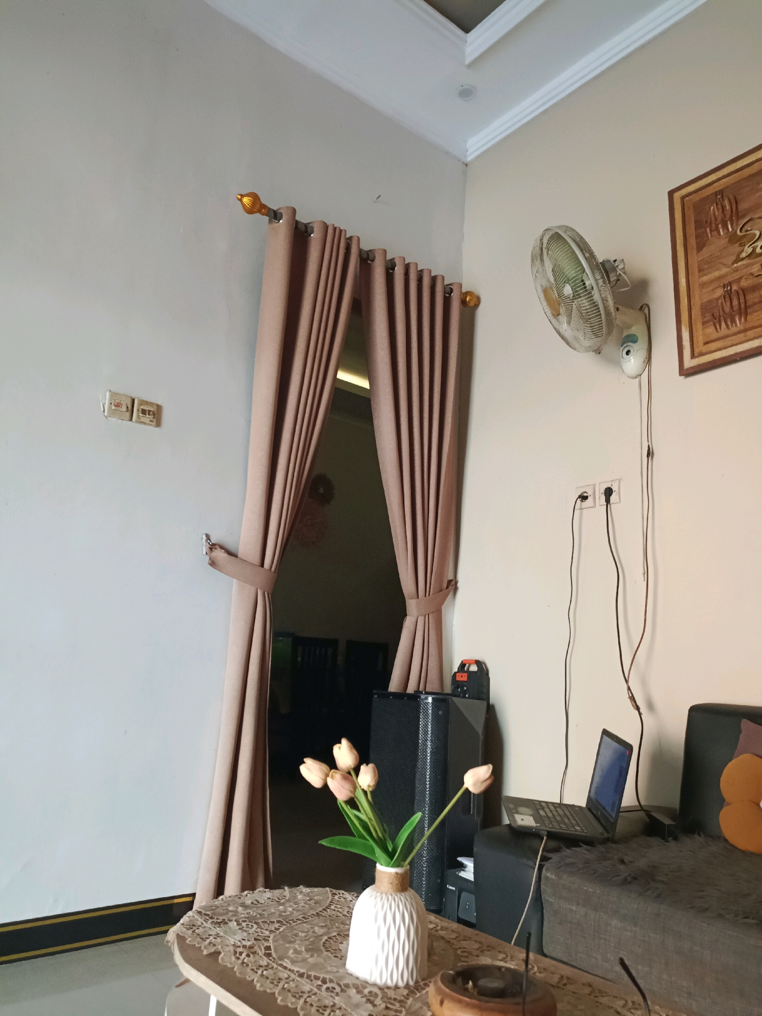 The image shows an indoor scene in what appears to be a living room or lounge area. The walls are a light neutral color, and the ceiling has white crown molding. On the left wall, there are two electrical outlets. A light brown curtain is drawn back with a tie-back, revealing a doorway with a dark interior.
Mounted on the right wall is a white electric fan. To the right of the fan is a framed piece of artwork, possibly religious in nature, with brown tones. Below the fan, there are two electrical outlets with black cables plugged into them.
In the foreground, a circular coffee table is covered with a decorative lace tablecloth. On the table, there is a white vase with a bunch of light pink tulips and a brown ashtray.
Behind the coffee table, there is a large black speaker to the left, and to the right, a dark gray sofa with a furry texture. On the sofa, a laptop is placed on what appears to be a dark ottoman or cushion. An orange throw pillow with a circular design is visible on the right side of the sofa.
The overall impression is of a modestly furnished indoor space. The mention of Rancang Kencono, Indonesia, suggests the geographical context of this scene.