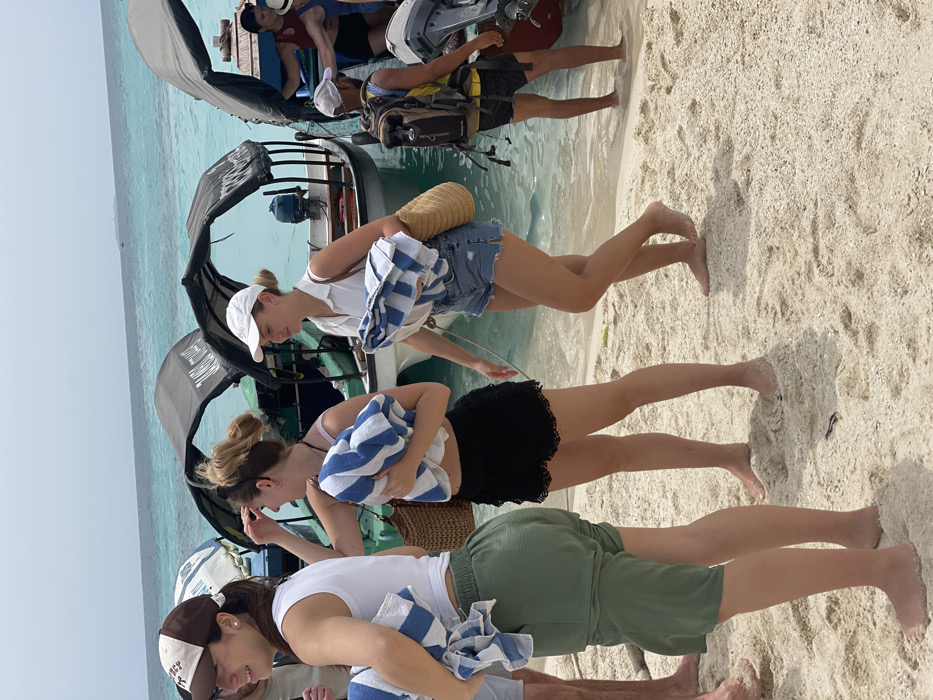 The image depicts a beach scene in Zanzibar, Tanzania, on a bright day. Several individuals are present on a sandy shore and in clear, shallow turquoise water, alongside multiple small boats.
In the foreground, three young adult females walk barefoot on light-colored sand, moving from left to right. The leftmost female, partially visible, wears a white tank top, olive green loose-fitting shorts, and a brown and white baseball cap. She carries a blue and white striped towel. The central female wears a light-colored top, black lace-trimmed shorts, and has her hair tied back. She holds a blue and white striped towel and a woven straw bag. The rightmost female wears a white collared short-sleeved shirt, blue denim shorts, and a white baseball cap, with her hair also tied back. She carries a blue and white striped towel and a woven straw bag.
To the right, further into the mid-ground, a male individual stands in the shallow water. He wears dark shorts, a light-colored baseball cap, and a large multi-colored backpack (beige, brown, black).
In the background, several small, open boats are partially submerged in the clear ocean water near the shore. These boats feature dark canopies (one has "TANSA..." visible text) and visible outboard motors, suggesting they are used for passenger transport. The water exhibits a distinct light blue to turquoise color, indicating a shallow, tropical environment. The sandy beach is composed of fine, light-colored sand with some shell fragments. The sky is clear, suggesting sunny conditions.