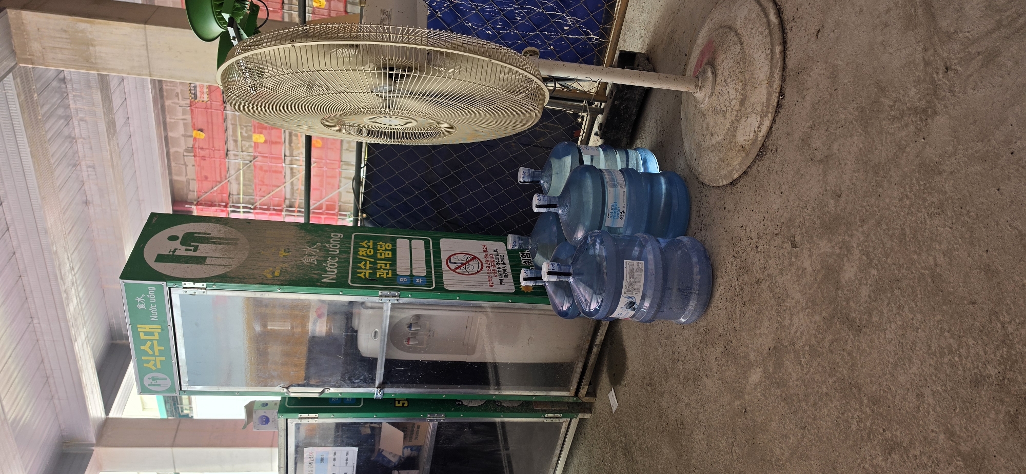 An outdoor scene captures a water cooler unit, a large oscillating fan, and several large water jugs. The water cooler, a white appliance with red and blue spigots, is housed within a green metal frame adorned with Korean and Vietnamese text and pictograms indicating a water dispenser. The text "식수대" and "Nước uống" translate to "Drinking water fountain" or "Water supply." To the right of the water cooler, a large, cream-colored oscillating fan stands on a circular base. In front of the water cooler and fan, four large, transparent blue plastic water jugs are arranged on the concrete ground. The jugs are sealed with white caps and some have labels. The background reveals construction scaffolding and netting, suggesting the location is a construction site. The ground surface is concrete, with some dirt and debris present. The overall setting appears to be a functional, temporary setup at a construction area in Pyeongtaek-si, South Korea.