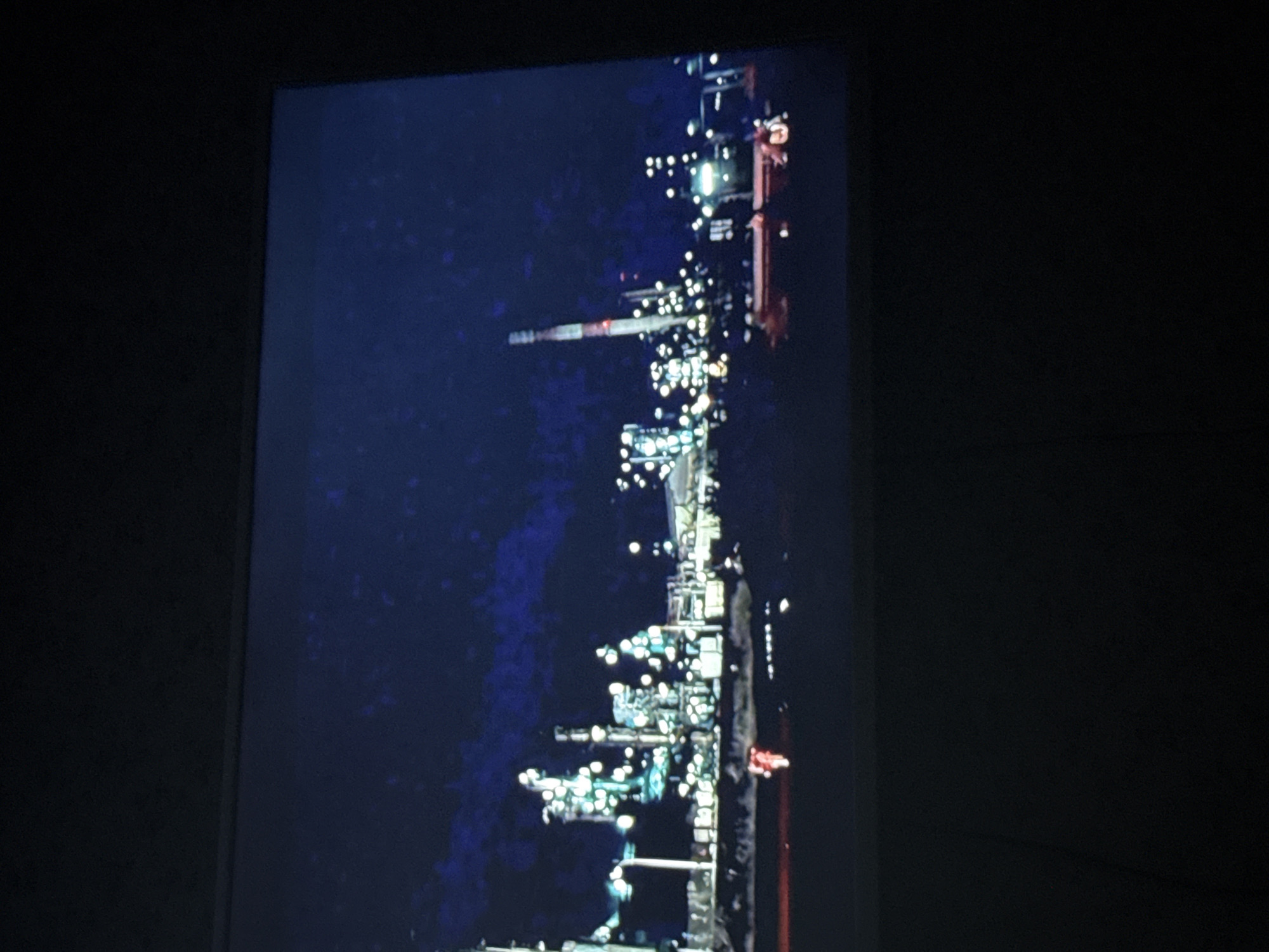 The image shows a nighttime view of an industrial facility, likely a refinery or a chemical plant, with numerous illuminated structures, towers, and pipes. A tall chimney or smokestack is visible in the center of the frame. The facility is adjacent to a dark body of water. The image seems to be taken from a distance, possibly from an elevated position. Without additional information, the specific location within Mytishchi, Russia cannot be determined.