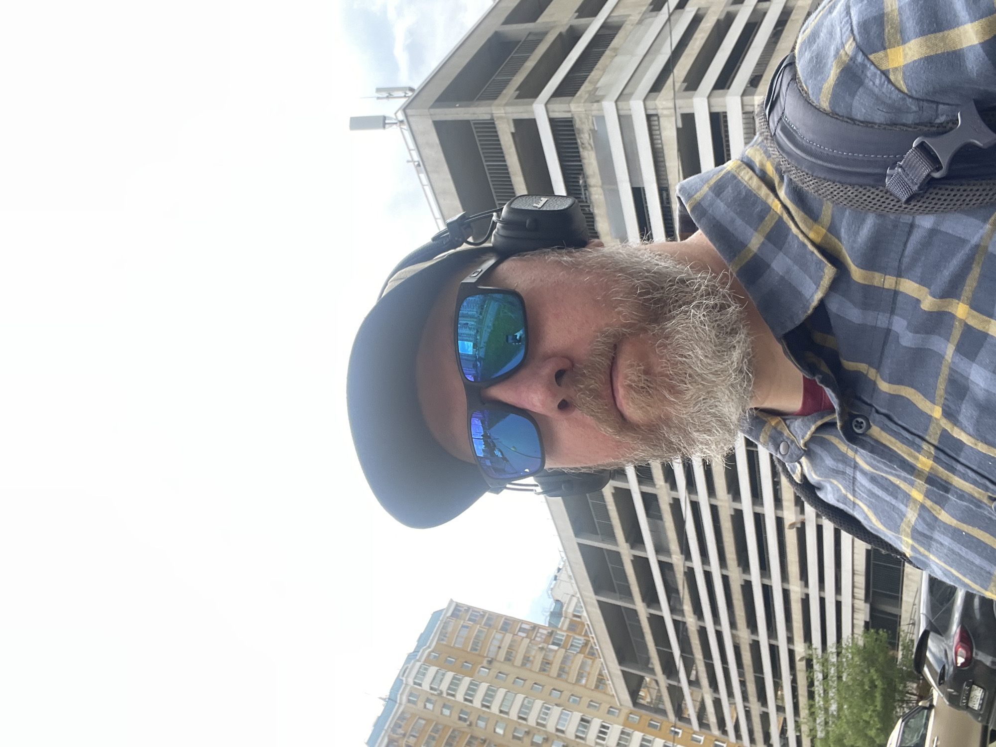 A selfie of a middle-aged man with a gray beard, wearing a blue and yellow plaid shirt, a dark baseball cap, and blue mirrored sunglasses. He is wearing headphones around his neck and has a backpack on. The buildings in the background are in Khimki, Russia. A multi-story parking garage with white vertical lines and a building with many windows are visible. A black car and some greenery are also visible in the background. The sky is overcast.
