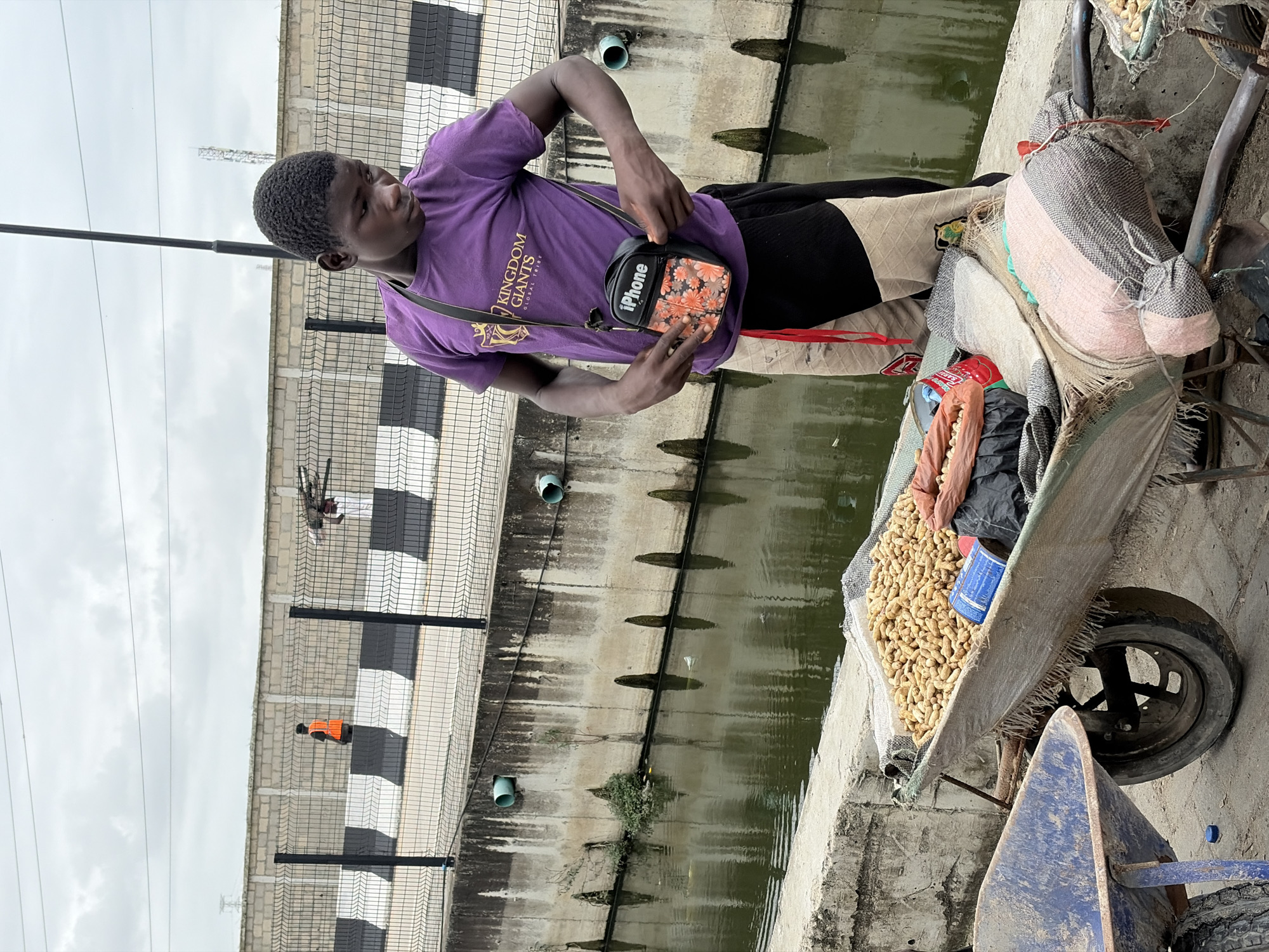 A young Black male, wearing a purple t-shirt with a "Kingdom Giants Global Tribe" graphic and a crossbody bag, stands next to a wheelbarrow filled with peanuts. He is adjusting his bag. The wheelbarrow is positioned on a paved surface next to a concrete embankment. In the background, a narrow canal with murky water runs alongside the embankment, which is bordered by a chain-link fence. Beyond the fence, a low concrete wall with black and white stripes is visible. In the distance, two individuals are walking, one wearing an orange vest. The sky is overcast. The scene is located in Igboefon, Nigeria.