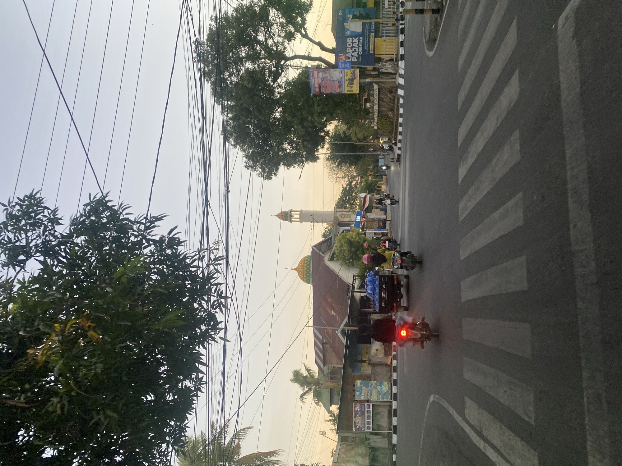 The image depicts a street scene in Bantaeng, Indonesia, likely near a significant intersection. In the foreground, a pedestrian crossing is clearly visible on the asphalt road. Several motorcycles are in motion. One motorcycle, ridden by a person wearing a black jacket and white pants, is in the left lane, its red taillight illuminated. Another motorcycle, with a rider wearing a yellow jacket and pink helmet, is to the right. Behind them, a small utility truck carries several large blue containers, possibly water bottles.
The background reveals a mosque with a distinctive green and yellow dome, and a tall, slender minaret. Adjacent to the mosque are buildings with brown, sloping roofs and some brick structures. Trees with lush green foliage are present, including a large tree to the right of the frame and branches of another tree in the upper left corner. Overhead, a complex network of power lines stretches across the sky, which is illuminated with the soft light of dusk or dawn, exhibiting hues of pale orange and yellow. Several billboards and signs are visible on the side of the road, including one that appears to advertise tax services. The overall impression is of a moderately busy urban environment during transitional light conditions.