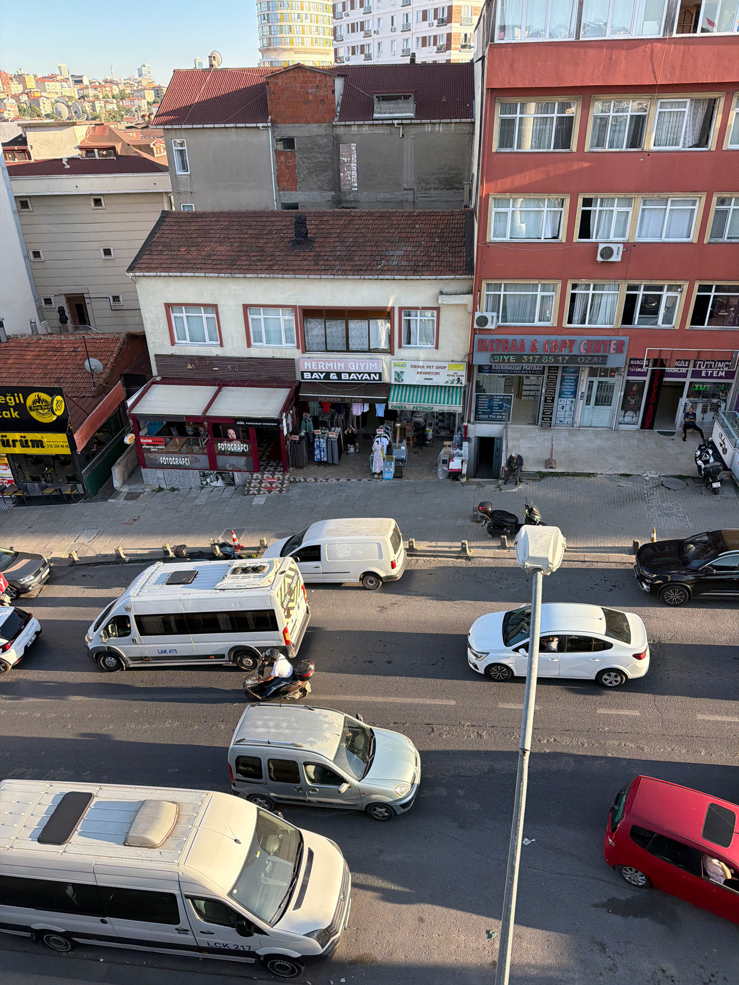 An elevated, long shot captures a street scene in Ataşehir, Turkey, under a clear, bright sky. The street is lined with a mix of commercial and residential buildings, featuring a variety of architectural styles and roof types, including red tile and flat roofs. Several vehicles are present on the road, including a white minibus with "LAK 475" visible on its side, a white sedan, a silver minivan, and a red hatchback. A person is seen riding a motorcycle. The ground level of the buildings hosts various businesses, indicated by signage such as "MATBAA & COPY CENTER", "NERMİN GİYİM BAY & BAYAN", and "FOTOGRAFCI". Several individuals are visible on the sidewalks and entering/exiting shops. Sunlight casts long shadows across the scene, suggesting it is either early morning or late afternoon.