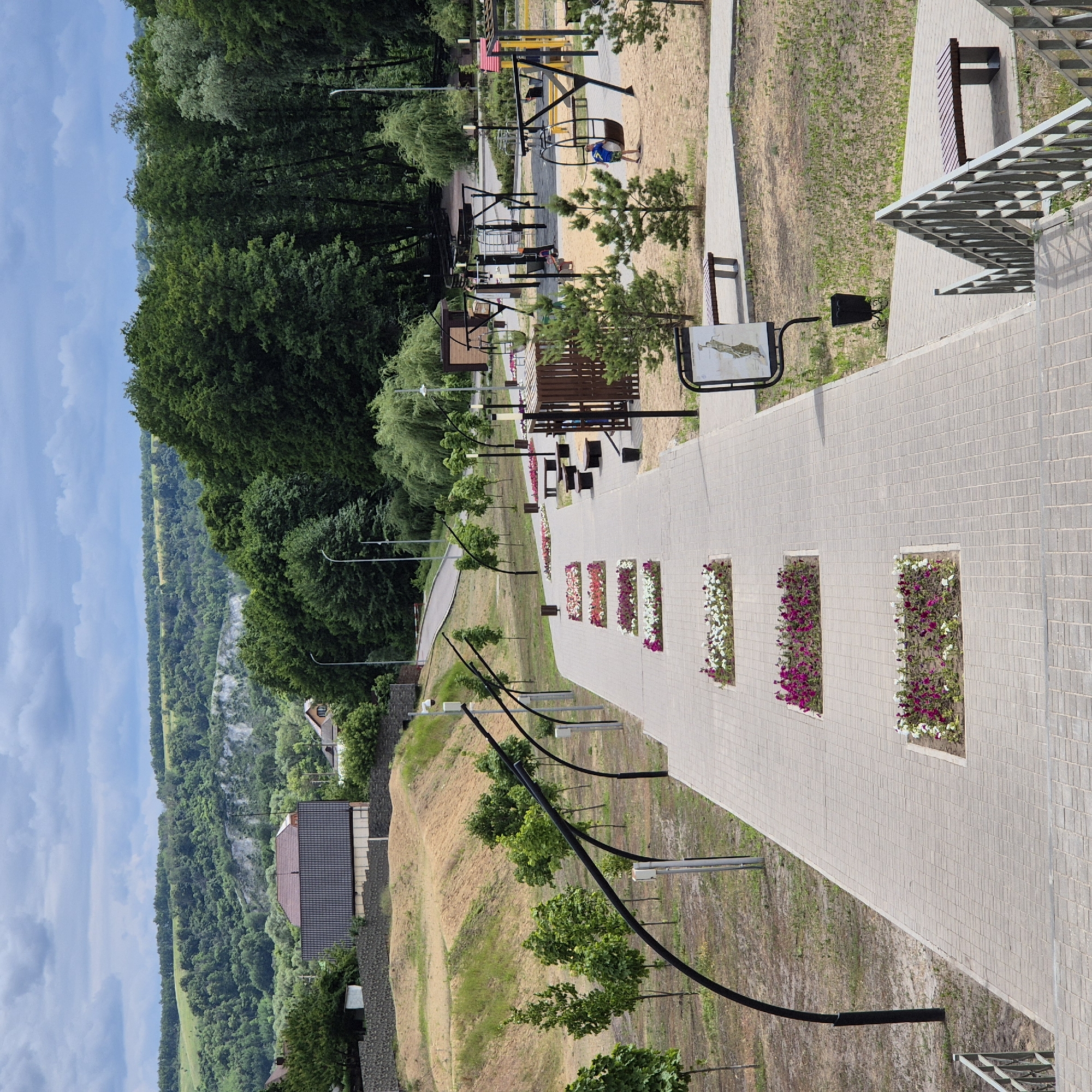 This is a scene from the city of Liski, Russia. The image captures a park area with a paved walkway lined by flower beds and small trees. In the background, there are houses, a playground with swings and climbing structures, and a dense forest covering rolling hills. The sky is overcast with clouds. The walkway leads towards the playground and the wooded hills. There are benches and trash cans placed along the walkway for public use. The scene appears to be a well-maintained public space designed for recreation and relaxation.