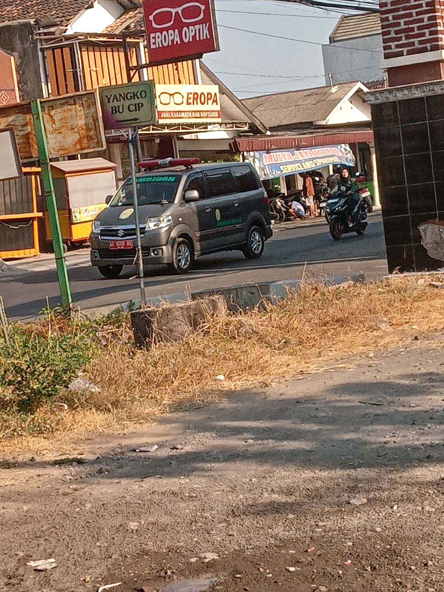The image shows a street scene in Banguntapan, Indonesia, on a bright, sunny day. In the foreground, dry grass and a dirt road are visible. A dark-colored utility vehicle with a license plate reading "AD 9006 BII" is parked on the asphalt road. The vehicle has a flashing red and blue light on its roof and text on its side that appears to say "JAMBIDAN." To the left of the vehicle, a green pole is partially visible, with a sign above it reading "YANGKO BU CIP" and an arrow pointing right. Further back, there are shops, including one with a red sign that says "EROPA OPTIK" and another with glasses displayed and text that reads "JUAN KACAMATA DAN SOFTLENS." Several motorcycles are parked near the shops, and one person is seated on a motorcycle, appearing to operate it. Another person is visible in the background near the motorcycles, possibly working on one. The overall impression is a busy commercial street.