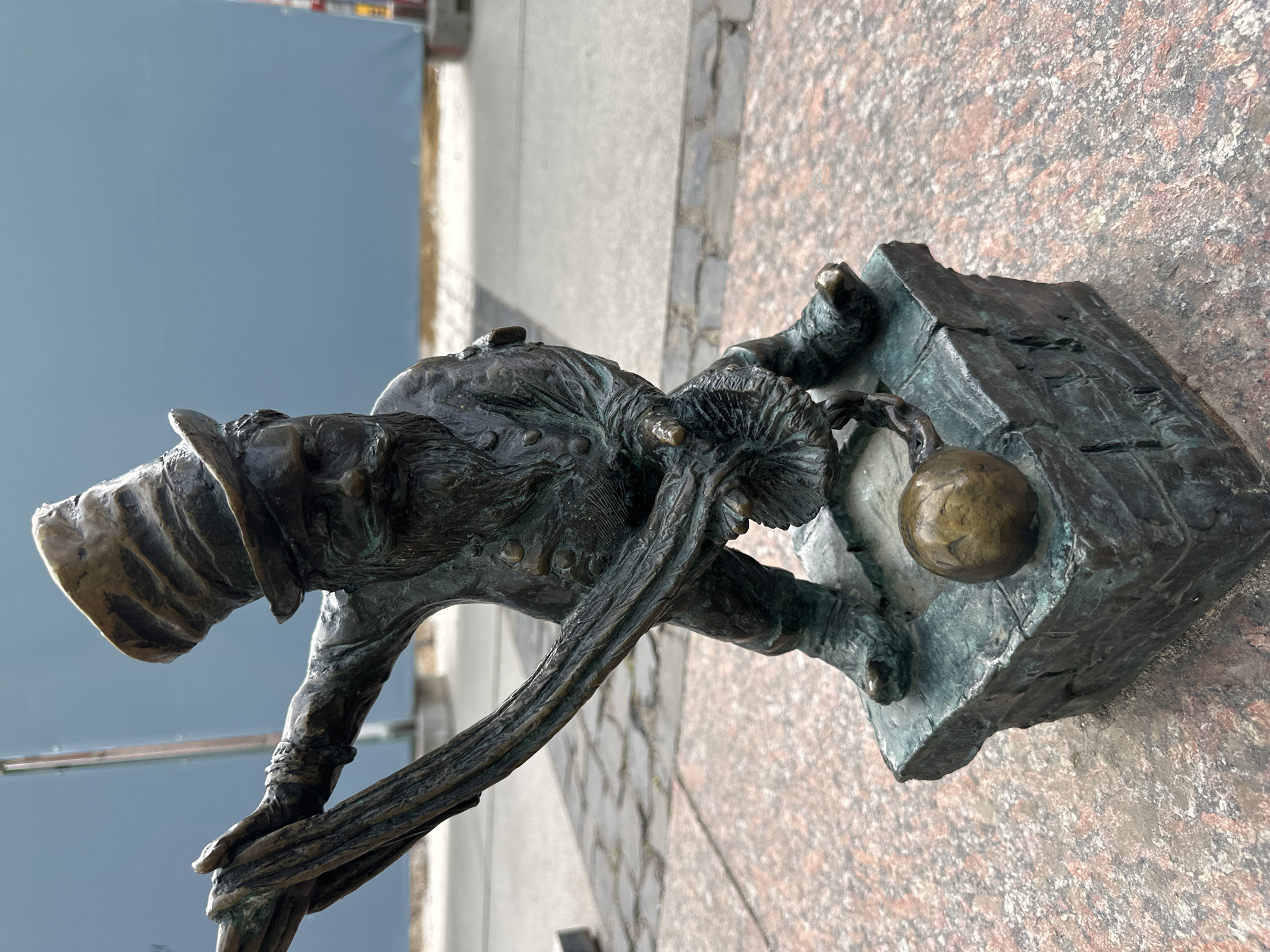 A bronze statue of a Wrocław dwarf is shown in an outdoor setting. The dwarf wears a hat, jacket and carries a sack or bag over its shoulder. It is bending forward. It stands next to a depiction of a brick building or wall with a gold-colored ball or sphere in the top corner. The statue sits on a surface made of red granite or a similar material. In the background is a sidewalk/street and a blue wall. The image is shot at an angle, with the background slightly blurred.