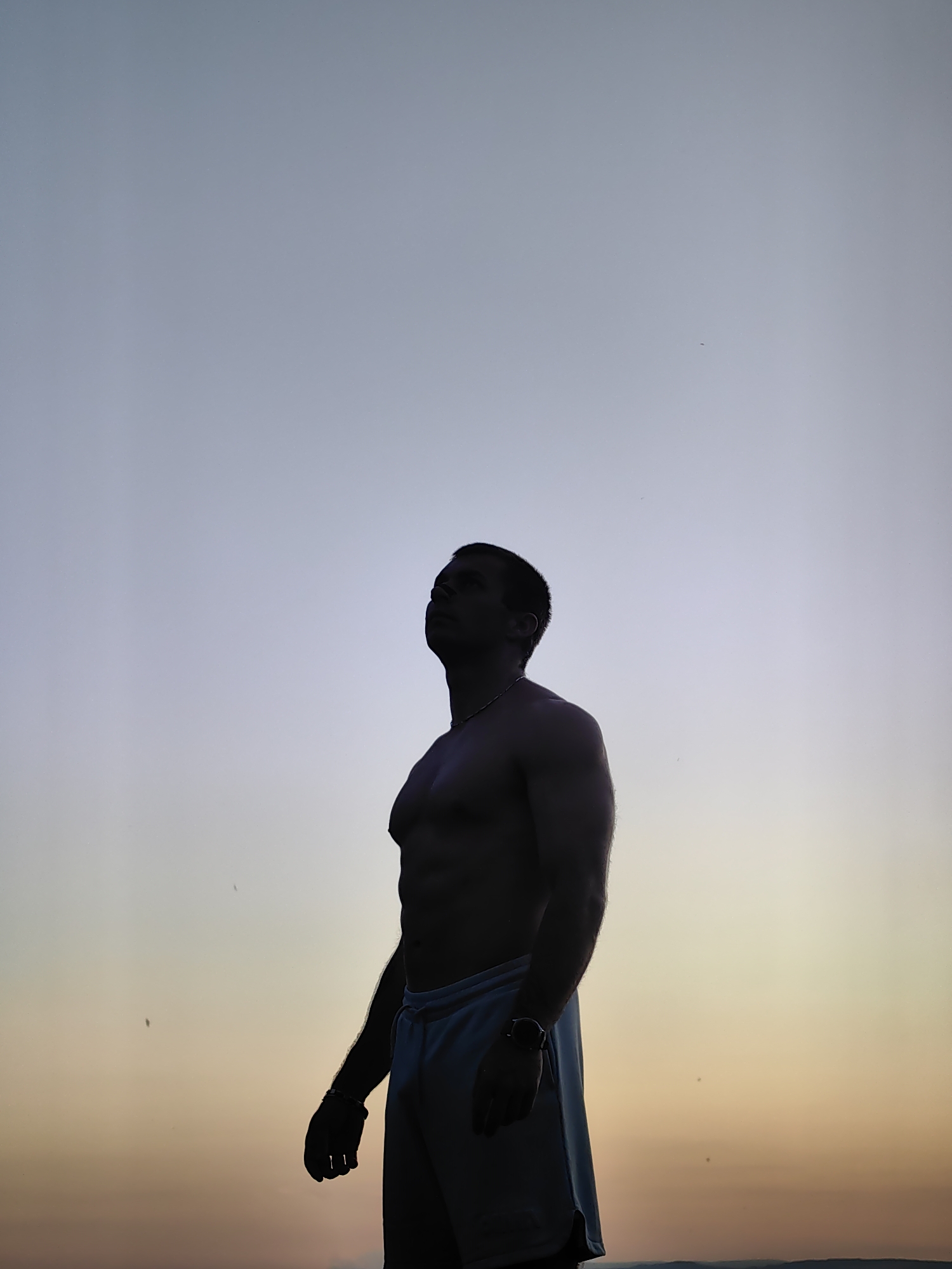 A full-shot silhouette of a muscular, shirtless man is captured against a gradient sky. The man is looking upwards, his head tilted back, with his arms hanging loosely at his sides. He is wearing light-colored shorts and a watch on his left wrist. The sky transitions from a pale blue at the top to a soft yellow and orange hue towards the horizon, suggesting either sunrise or sunset. The background is mostly devoid of detail, with only a faint hint of a distant landscape at the very bottom. There are a few small specks, possibly insects or dust, visible in the sky. This image is taken outdoors, likely in an open area, and given the information, the location in Varna, Bulgaria is the Black Sea coast, possibly near a beach or promenade, during twilight hours.