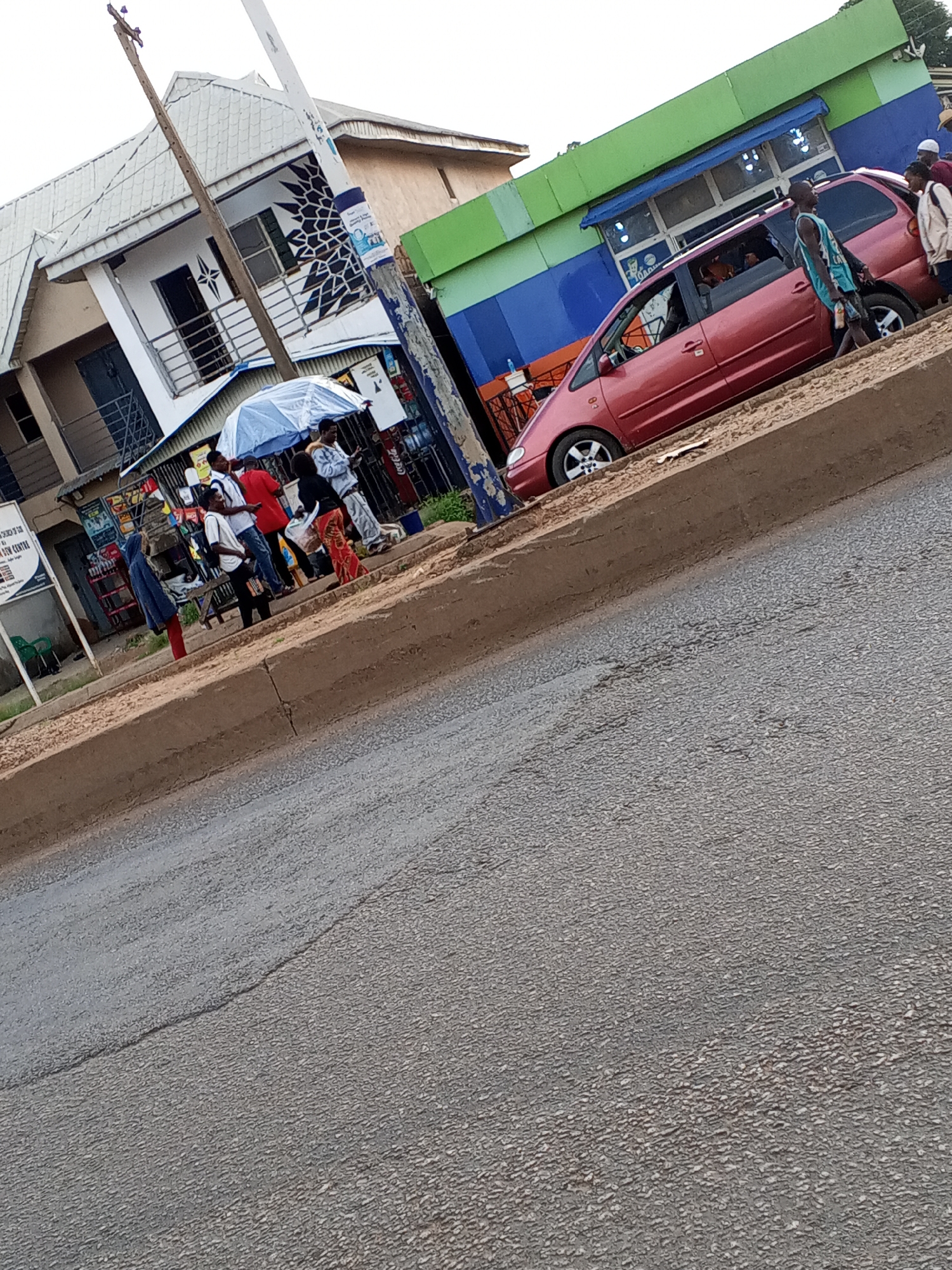 This is a wide, slightly tilted shot of a street scene in Bukuru, Nigeria. On the left, a light-colored building with dark trim and a balcony sits next to a wooden utility pole. A blue and white striped sign is visible on the pole. In front of the building, a group of people are gathered under a large, light blue umbrella, with some standing and others appearing to be sitting on a bench. A variety of items, possibly goods for sale, are displayed on shelves to the right of the umbrella.
Further to the right, a red minivan is parked at the edge of a sidewalk. Several people are near the minivan; one individual, a young man wearing a light blue tank top and shorts, is walking towards the viewer's right. Another person, wearing a white top and dark pants, is standing by the minivan's open rear door. A woman in a red and black patterned skirt is also visible, holding a white bag. Behind the minivan, a building with a green and blue facade is visible, with what appears to be a storefront. The street in the foreground is paved and shows signs of wear. The overall lighting suggests it is daytime, possibly late afternoon.