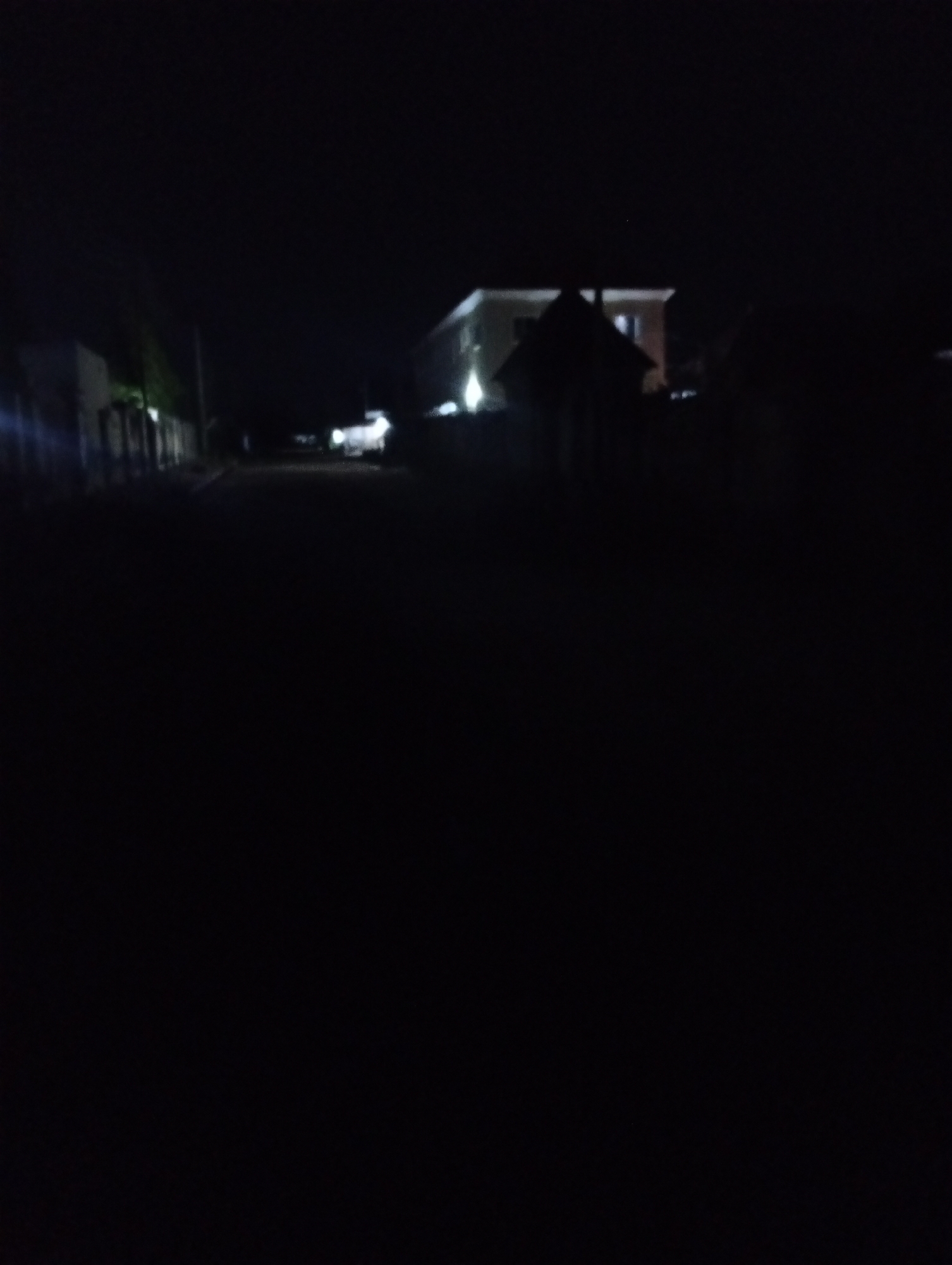 This image depicts a night scene in Adankolo, Nigeria. A dark road stretches into the distance, flanked by buildings and fences on either side. In the distance, a two-story building with lit windows is visible. The overall environment is dimly lit, obscuring details in the foreground.