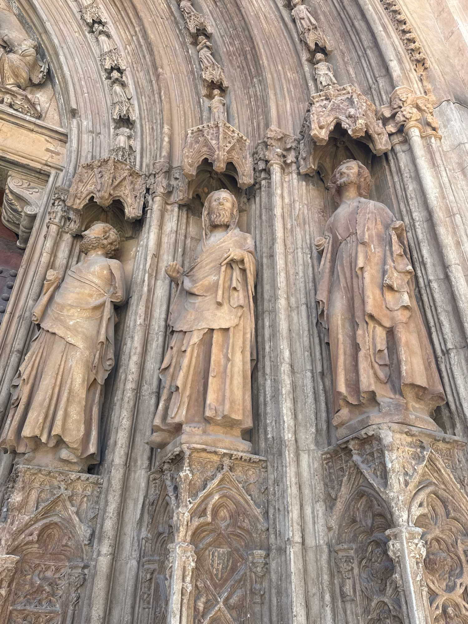 The image shows a detailed view of the exterior architecture of the Valencia Cathedral in Valencia, Spain. Specifically, it captures a section of the Gothic facade, focusing on three carved stone statues of men in robes, positioned within decorative niches. The statues are weathered and show signs of age, with intricate drapery and individualized facial features. They are set against a background of ribbed stone arches and decorative architectural elements, such as carved moldings and small figures. The lighting suggests it is daytime, with sunlight casting shadows that highlight the textures of the stone. The overall impression is one of ancient religious artistry and historical significance.